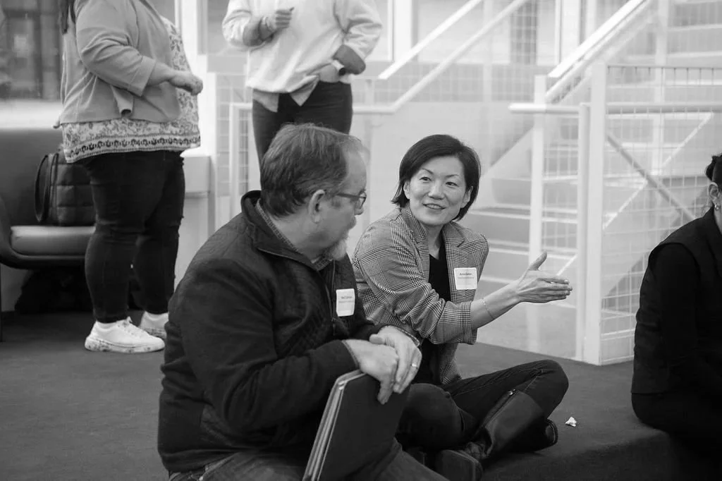 Two people sitting on the floor engaged in conversation, with one person gesturing as they speak. Others stand in the background in a modern indoor setting.