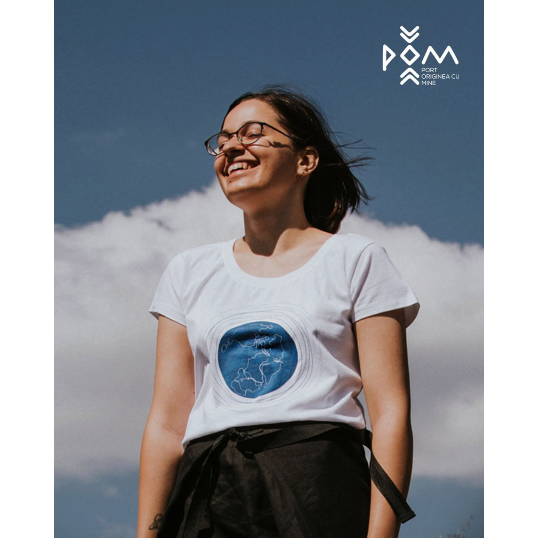 A woman smiling outdoors against a partly cloudy sky, wearing glasses, a white t-shirt with a blue circular design, and black pants tied at the waist. There is a logo and some text in the top right corner.