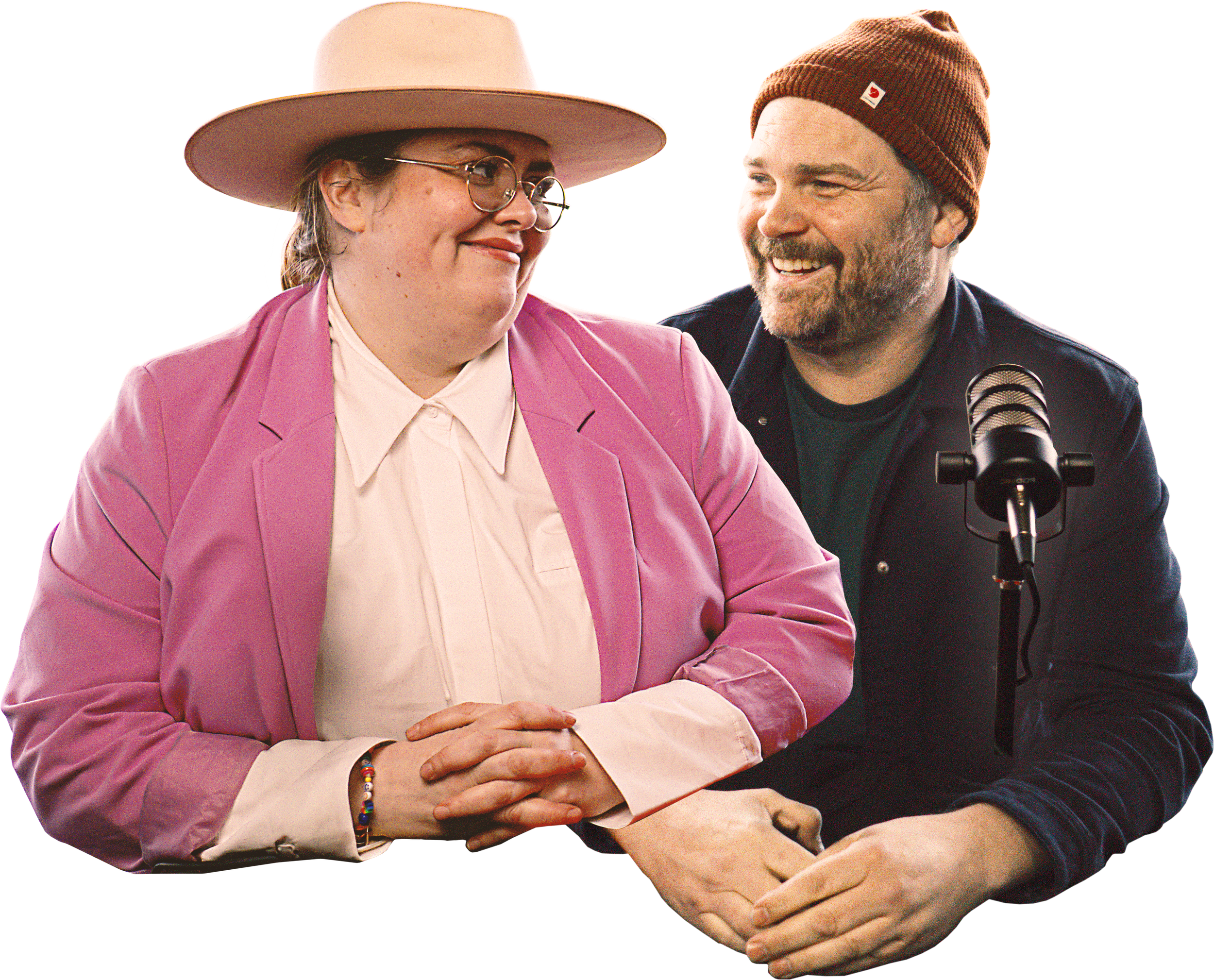 Two people sitting together, smiling, with a microphone nearby. One is a woman wearing a pink blazer, white blouse, large glasses, and a wide-brimmed hat, and the other is a man wearing a brown beanie, dark jacket, and a black shirt.