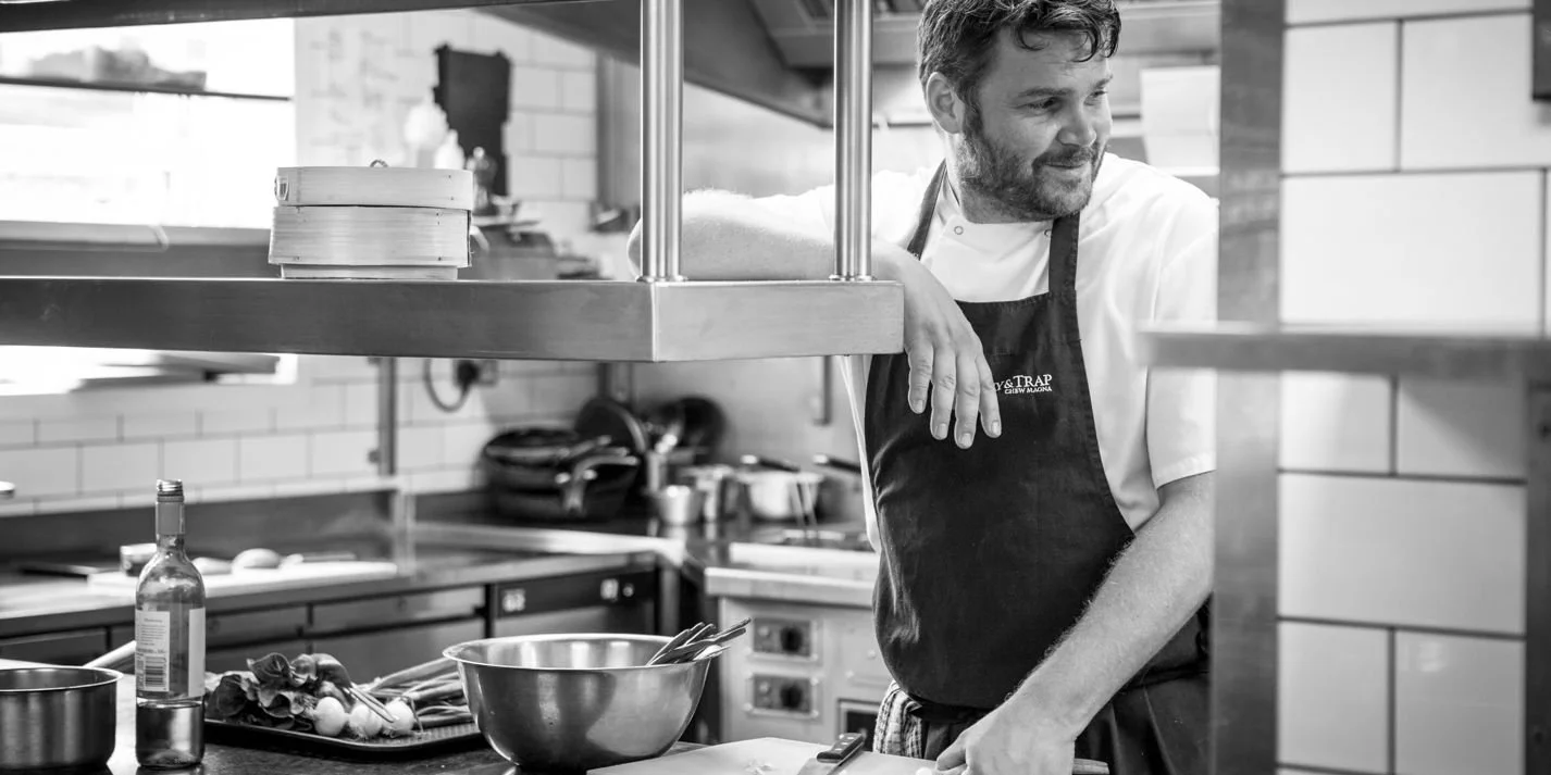 Josh Eggleton in a white chef's coat and black apron leaning on a kitchen counter in a restaurant kitchen, smiling and looking to the side, with kitchen utensils and ingredients on the counter.