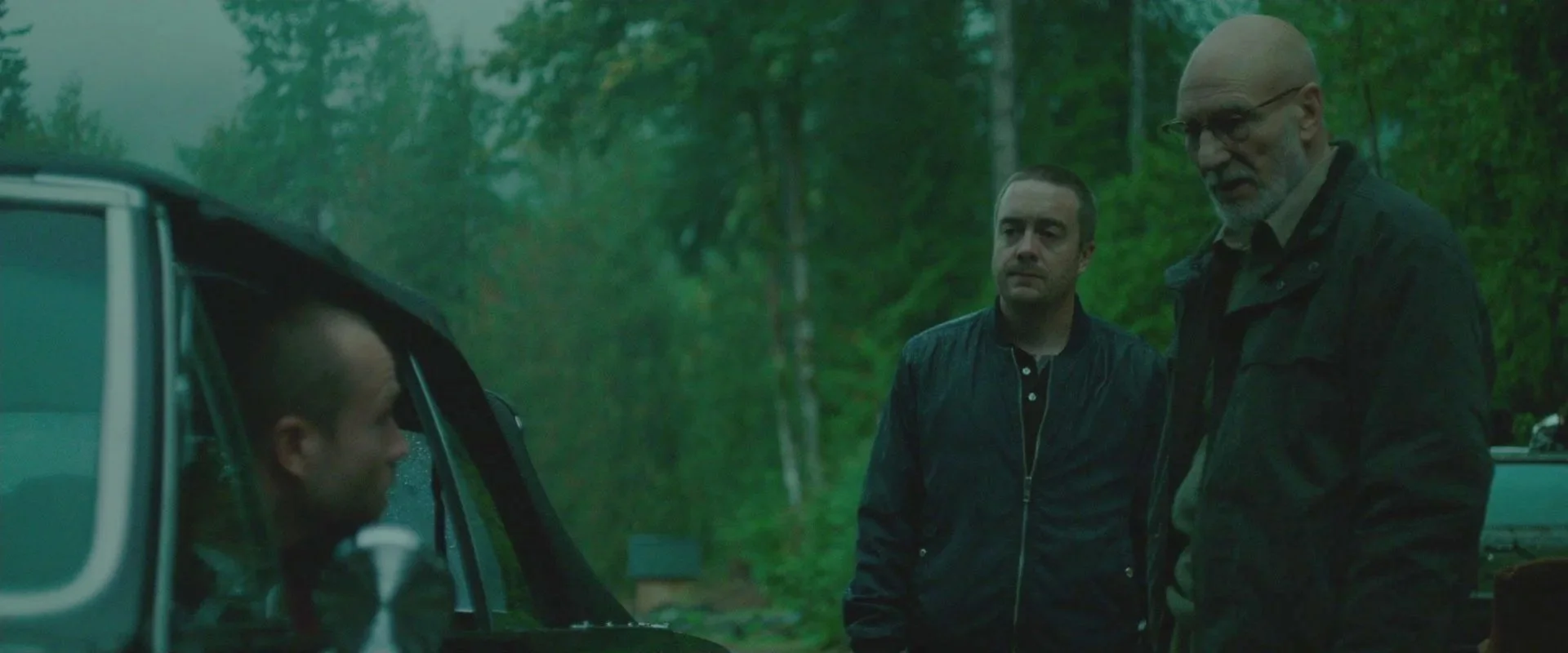 Three men in a wooded outdoor area, two standing and talking, one sitting inside a car with raindrops on the window.