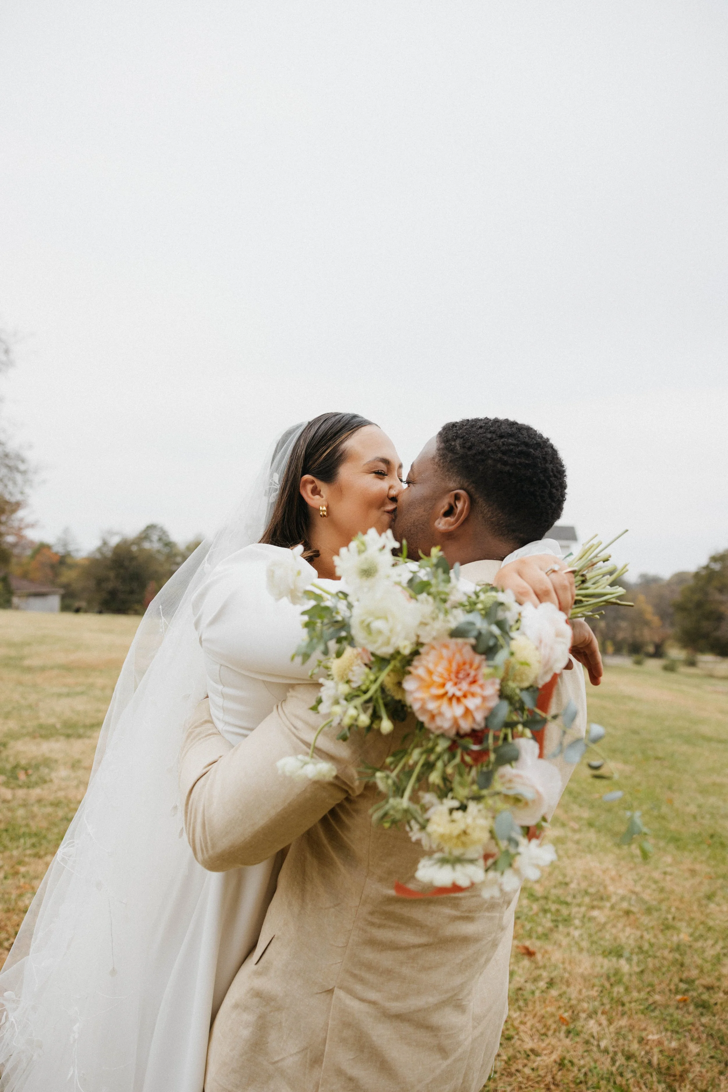 A newlywed couple sharing a kiss outdoors on a cloudy day, with the bride in a white gown and veil holding a bouquet of white, peach, and green flowers, and the groom in a light beige suit, embracing in a grassy area with trees in the background.