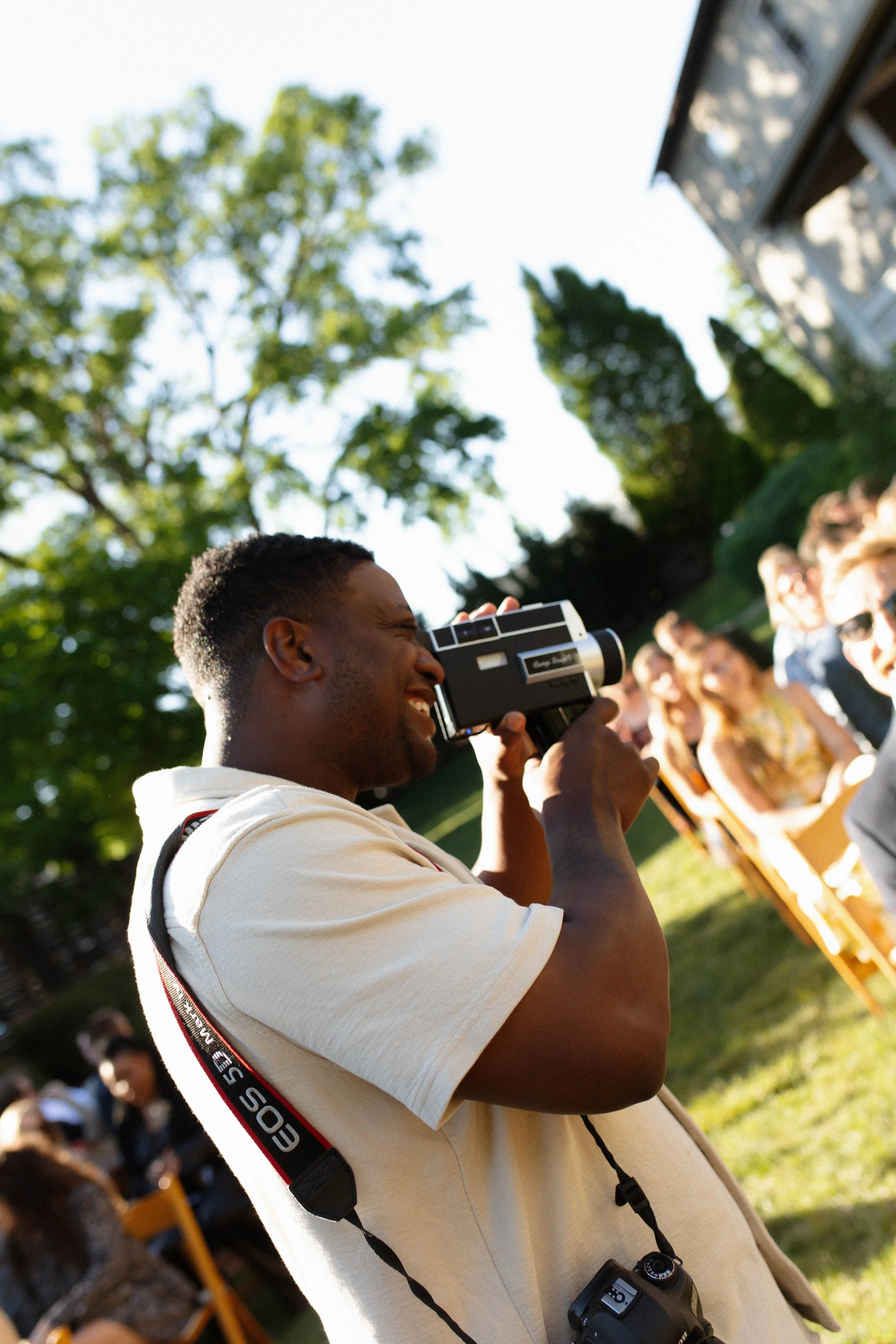 A young man with a camera around his neck enjoying an outdoor gathering, smiling and talking with people in the background on a sunny day with green trees and a building.