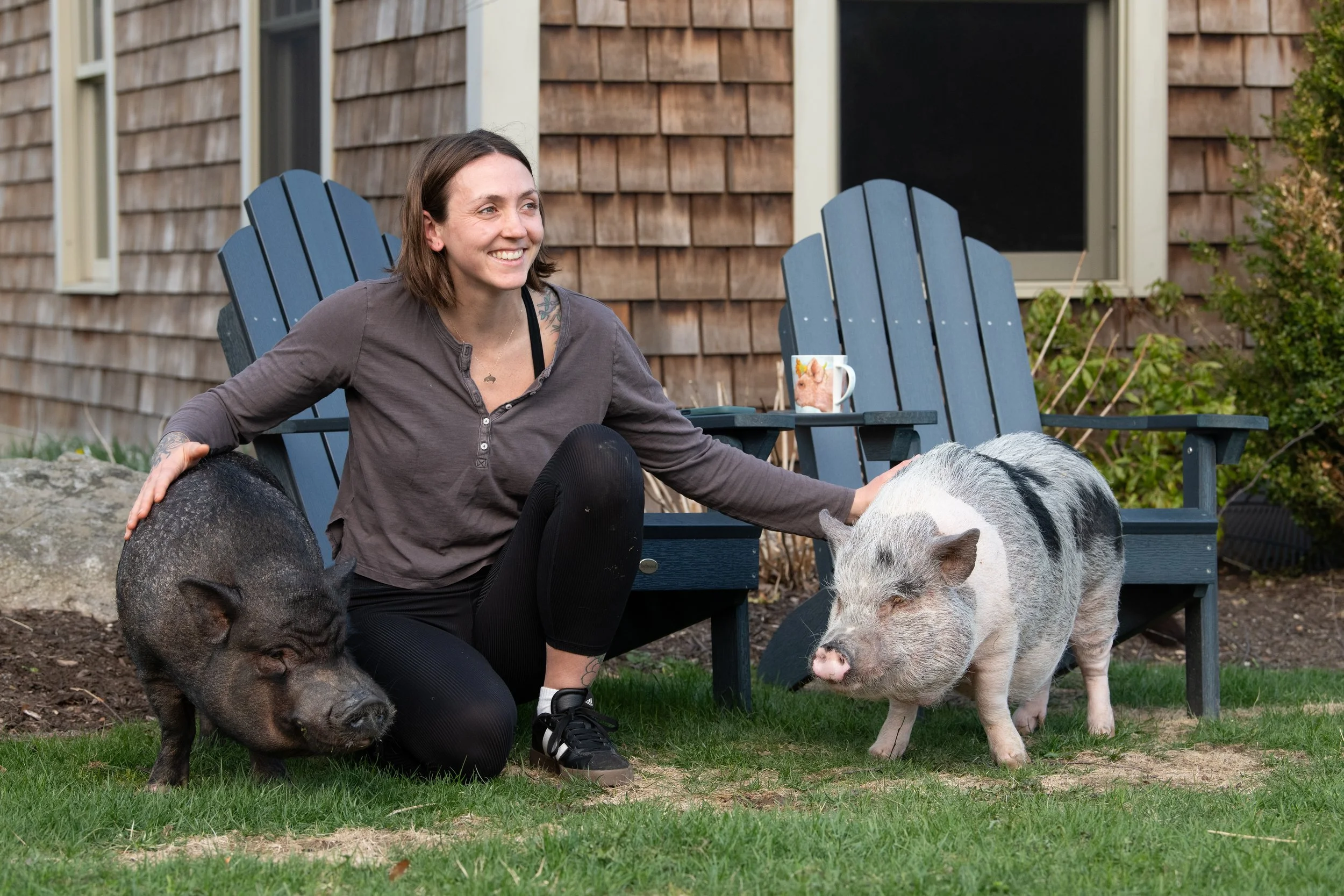 A woman with shoulder-length brown hair, smiling, sitting outdoors on green grass with a piglet and a pig, petting the piglet. She is in front of a wooden house with a window, on blue Adirondack chairs, near bushes.