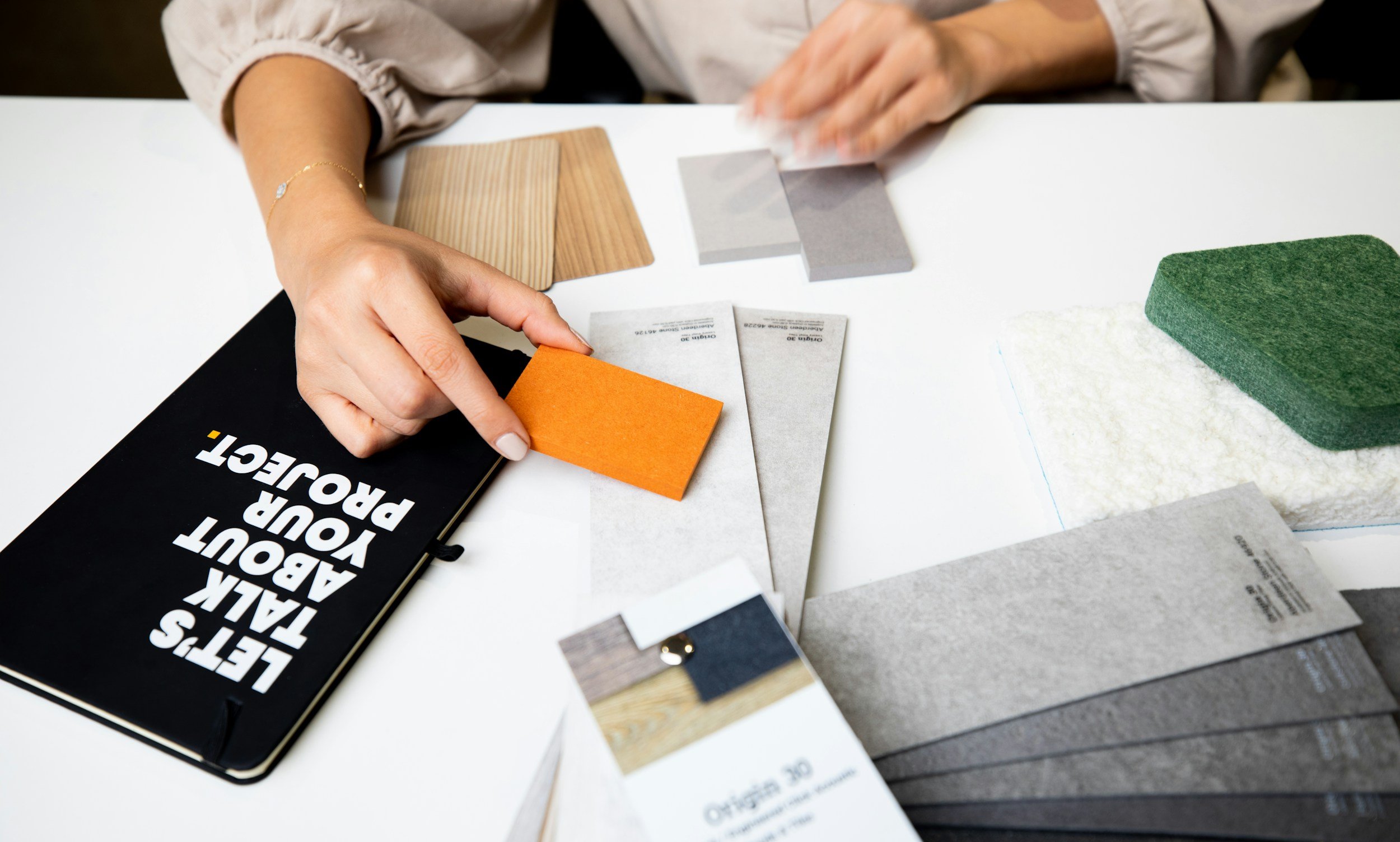 Close-up of a person's hand examining color and material samples laid out on a white table, with a black book titled 'Let's Talk About Your Project' nearby.