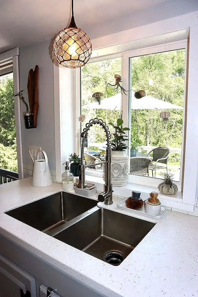 Kitchen sink area with a double basin stainless steel sink, a modern pull-down faucet, and various decorative plants and containers on the white countertop. Outside the window, there is a patio with outdoor furniture and umbrellas, surrounded by green trees.