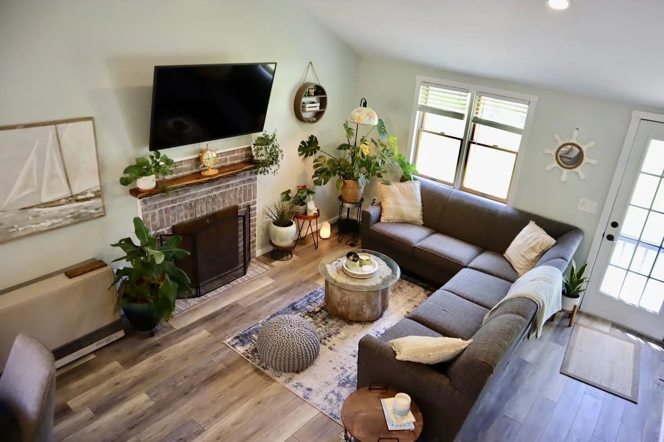 Living room with a gray sectional sofa, a round coffee table, a brick fireplace with a wall-mounted TV above it, potted plants, and windows letting in natural light.
