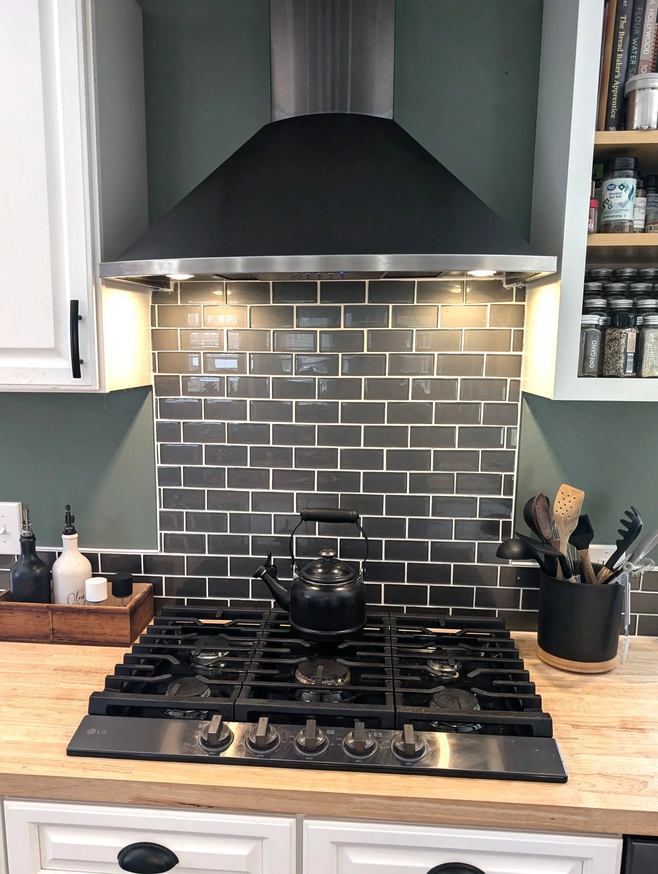 Kitchen stove with black kettle, black and white salt and pepper shakers, containers on a wooden countertop, and a black utensil holder with cooking tools. Gray subway tile backsplash. Range hood overhead.