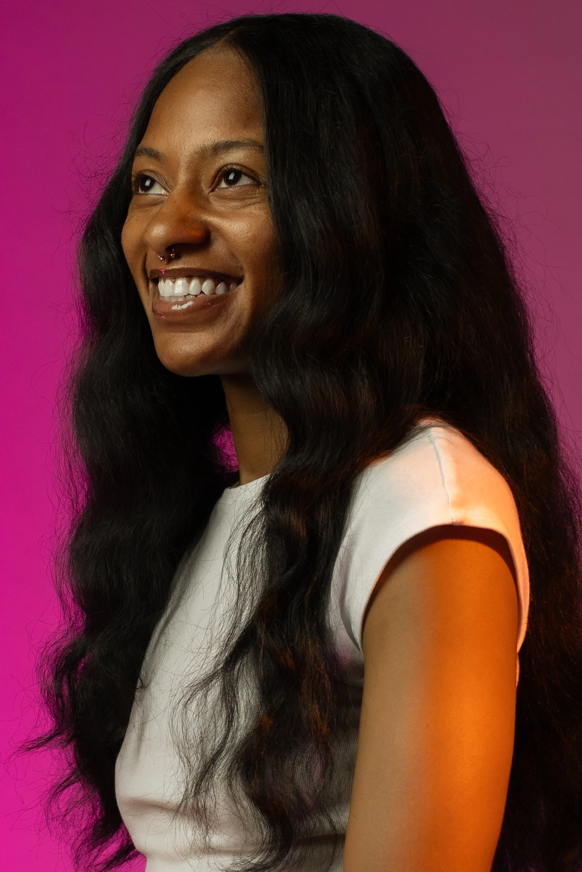 A woman with long, wavy black hair smiling against a pink and purple gradient background.