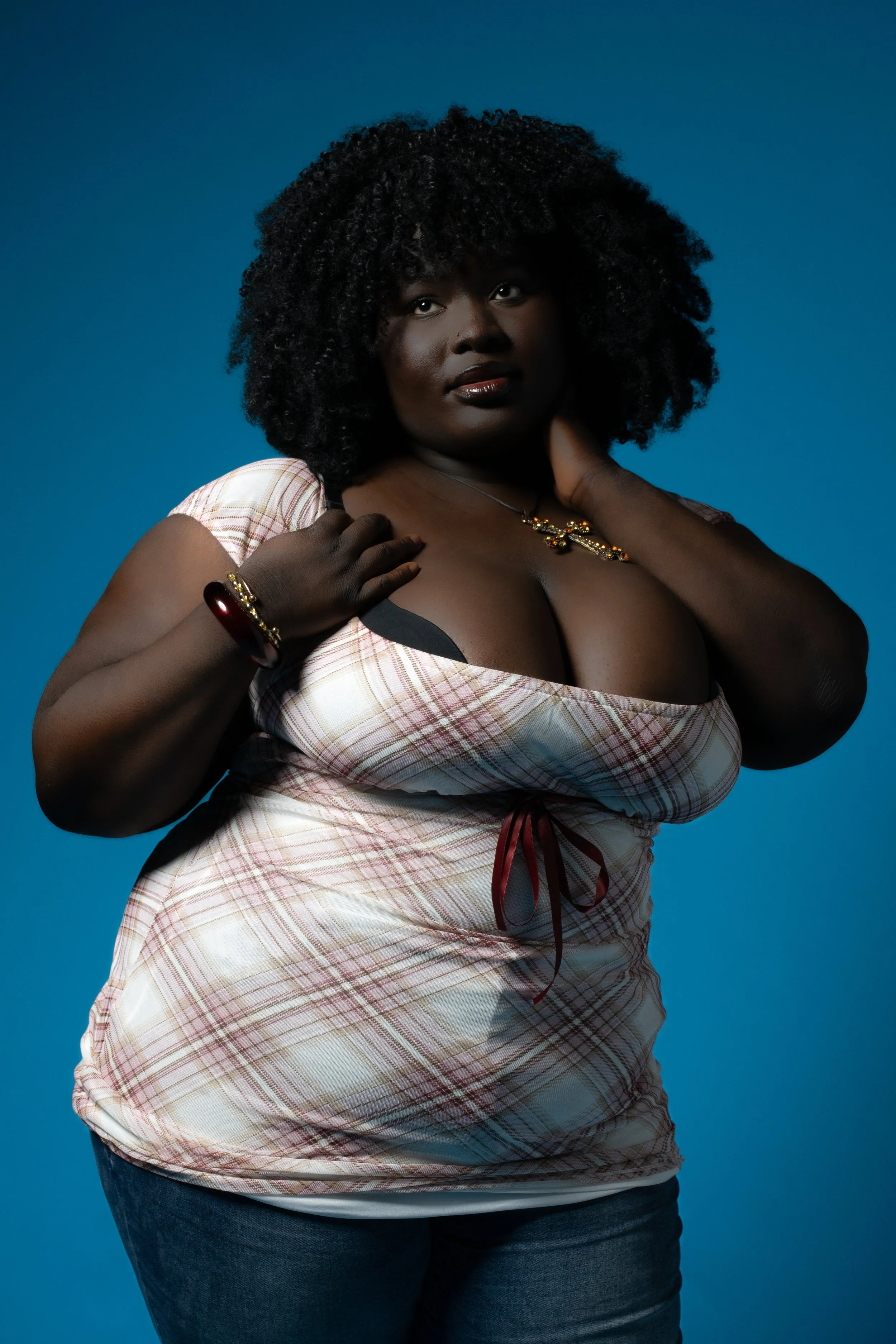 A woman with dark skin and curly black hair poses against a blue background, wearing a plaid top, jeans, gold jewelry, and a red bracelet.