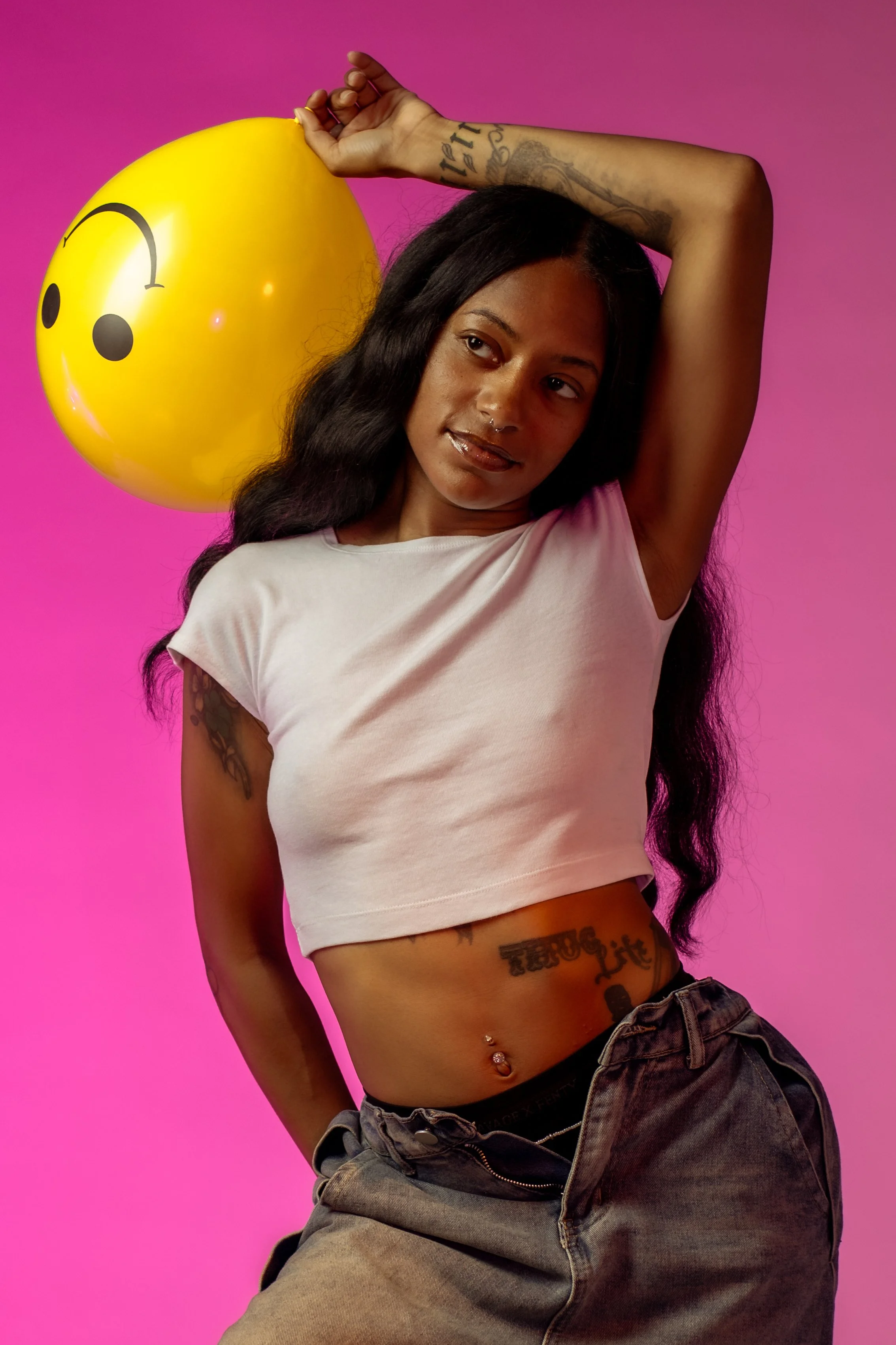Young woman with long black hair and tattoos, wearing a white crop top and gray pants, holding a yellow smiling face emoji balloon against a pink background.