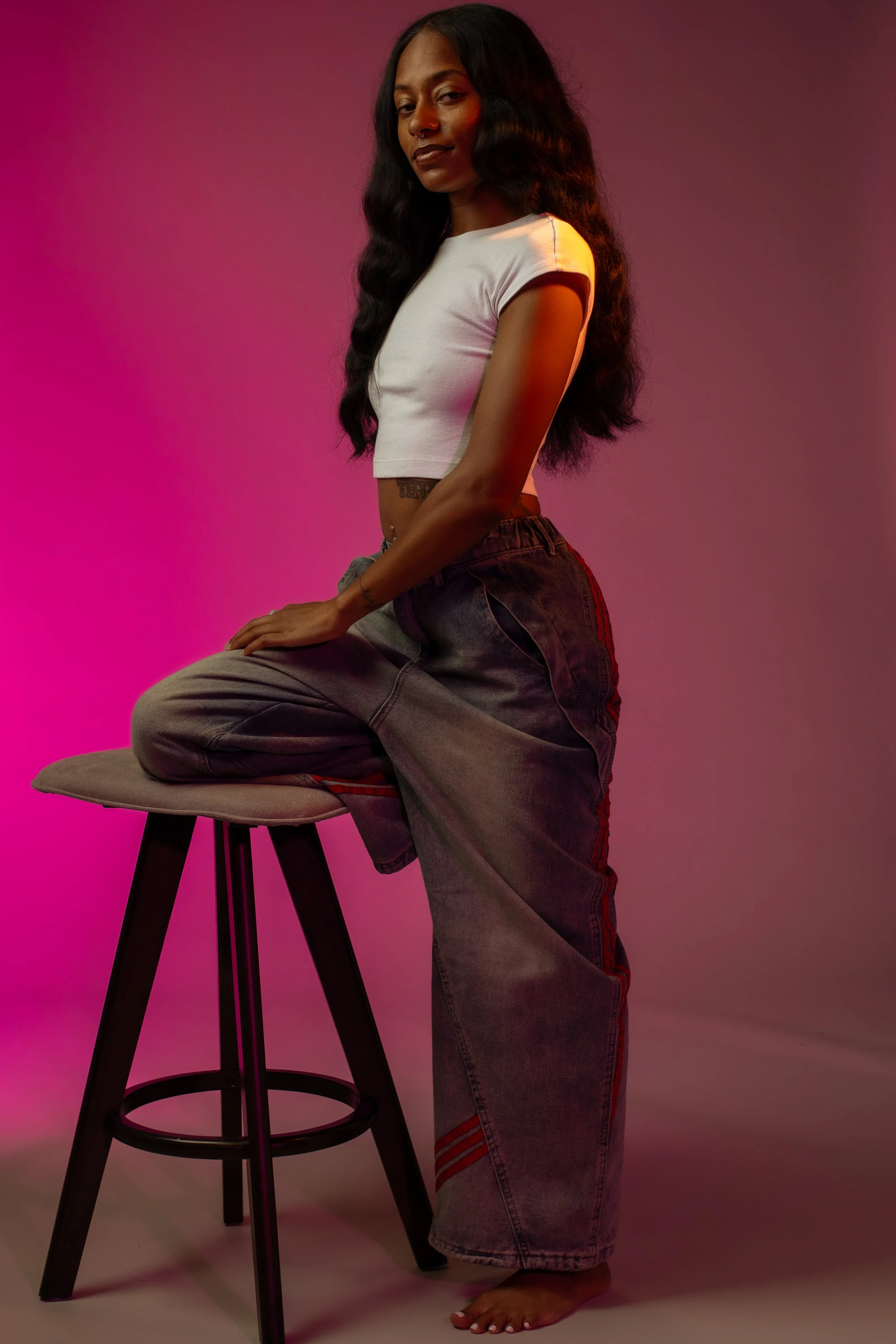 A young woman with long wavy hair and dark skin wearing a white cropped t-shirt and loose-fitting gray jeans, posing barefoot with her hand on a padded stool against a pink and gray gradient background.