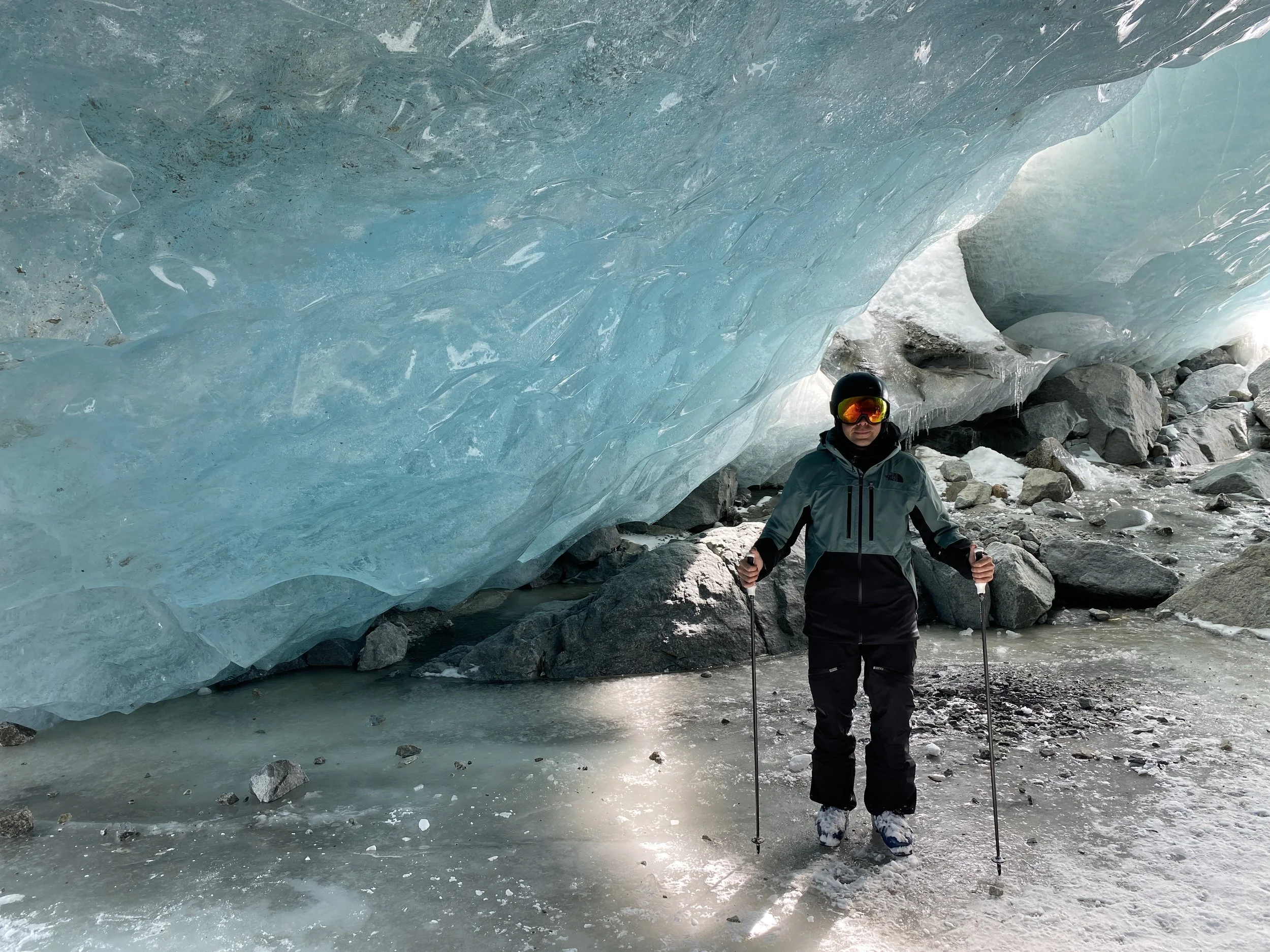 A person in winter gear, including a helmet and ski goggles, standing in front of an ice cave or glacier with icy walls and rocky terrain.