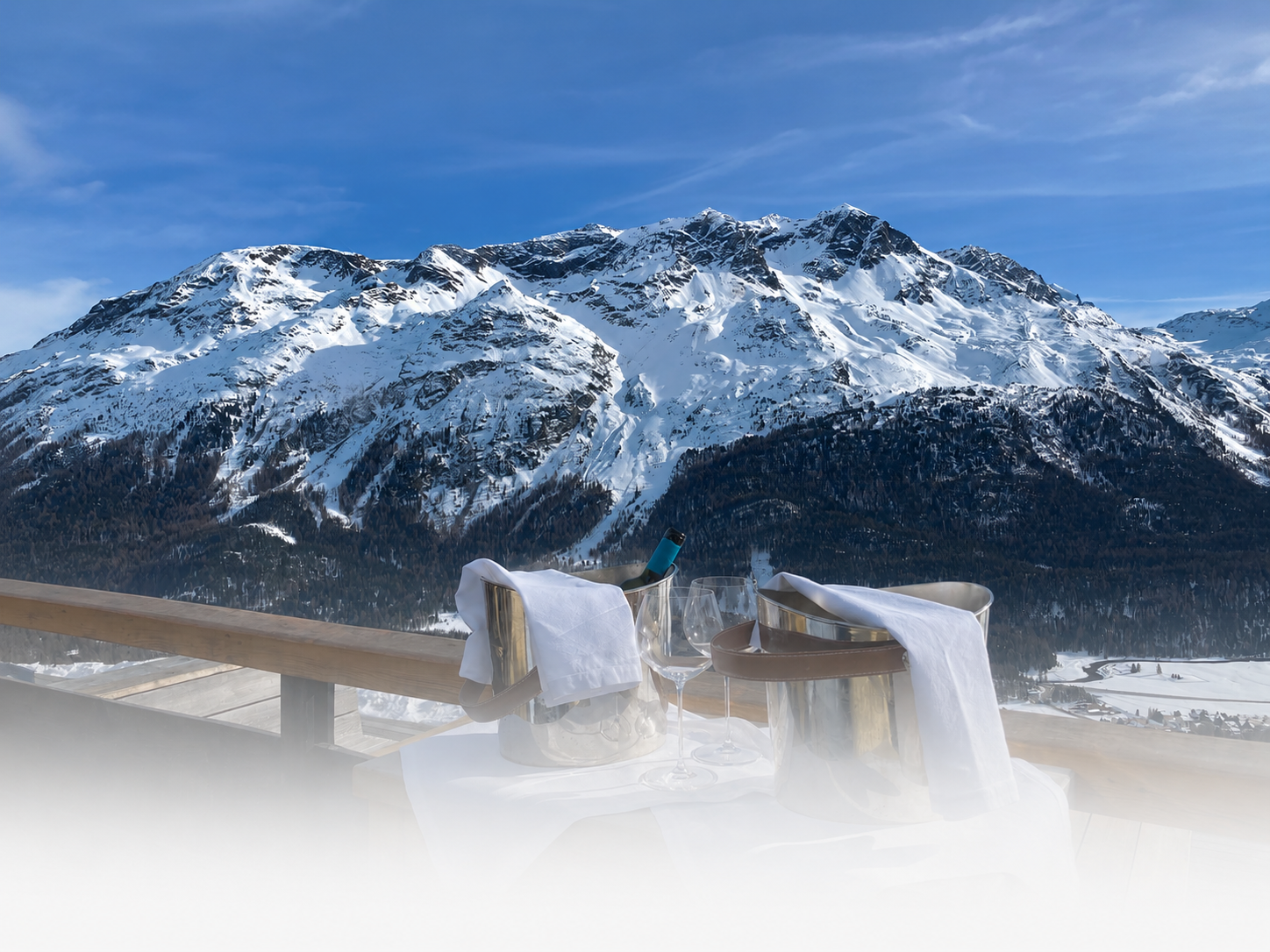A table on a wooden balcony with a bottle of wine, two filled wine glasses, and two wine buckets with towels, overlooking snow-covered mountains in the distance.