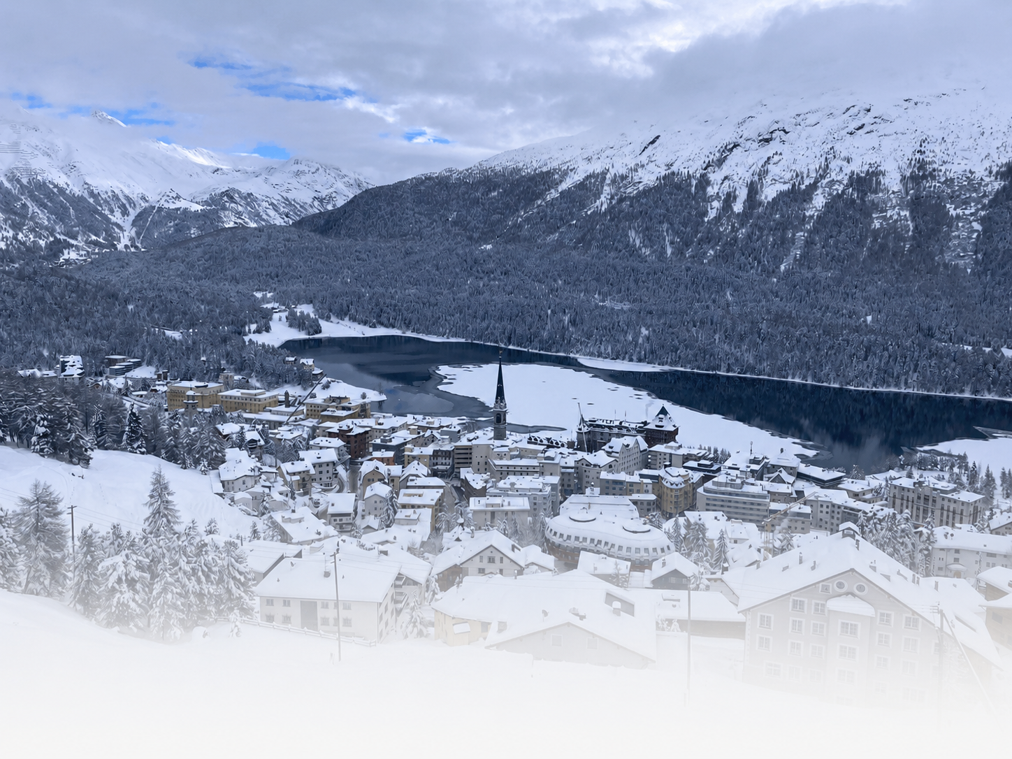 Snow-covered St. Moritz village with a lake, forest, and mountain range in the background.