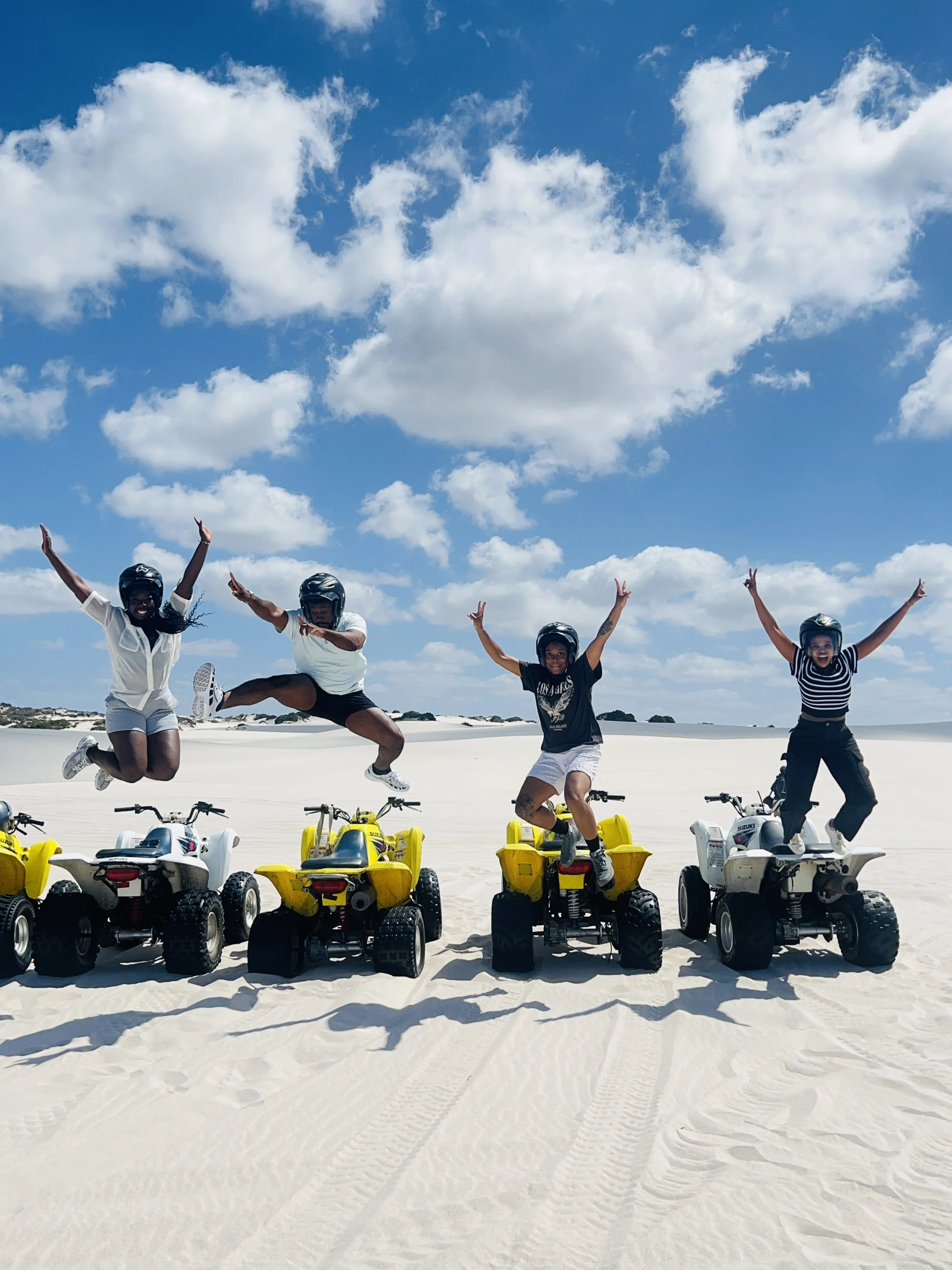 Four people wearing helmets jumping on mini quad bikes in a sandy desert under a blue sky with scattered clouds.