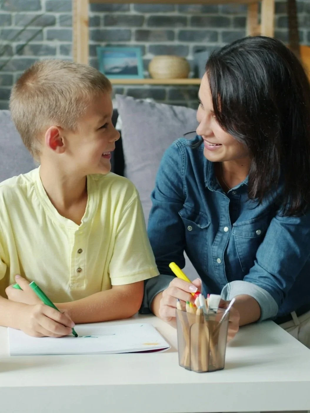 A woman and a young boy smiling at each other while sitting at a table, each holding a yellow highlighter, with a notebook and a glass container of pens and markers in front of them.