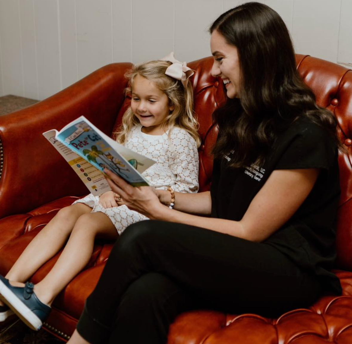 A woman and a young girl sitting on a red leather couch, reading a book together and smiling