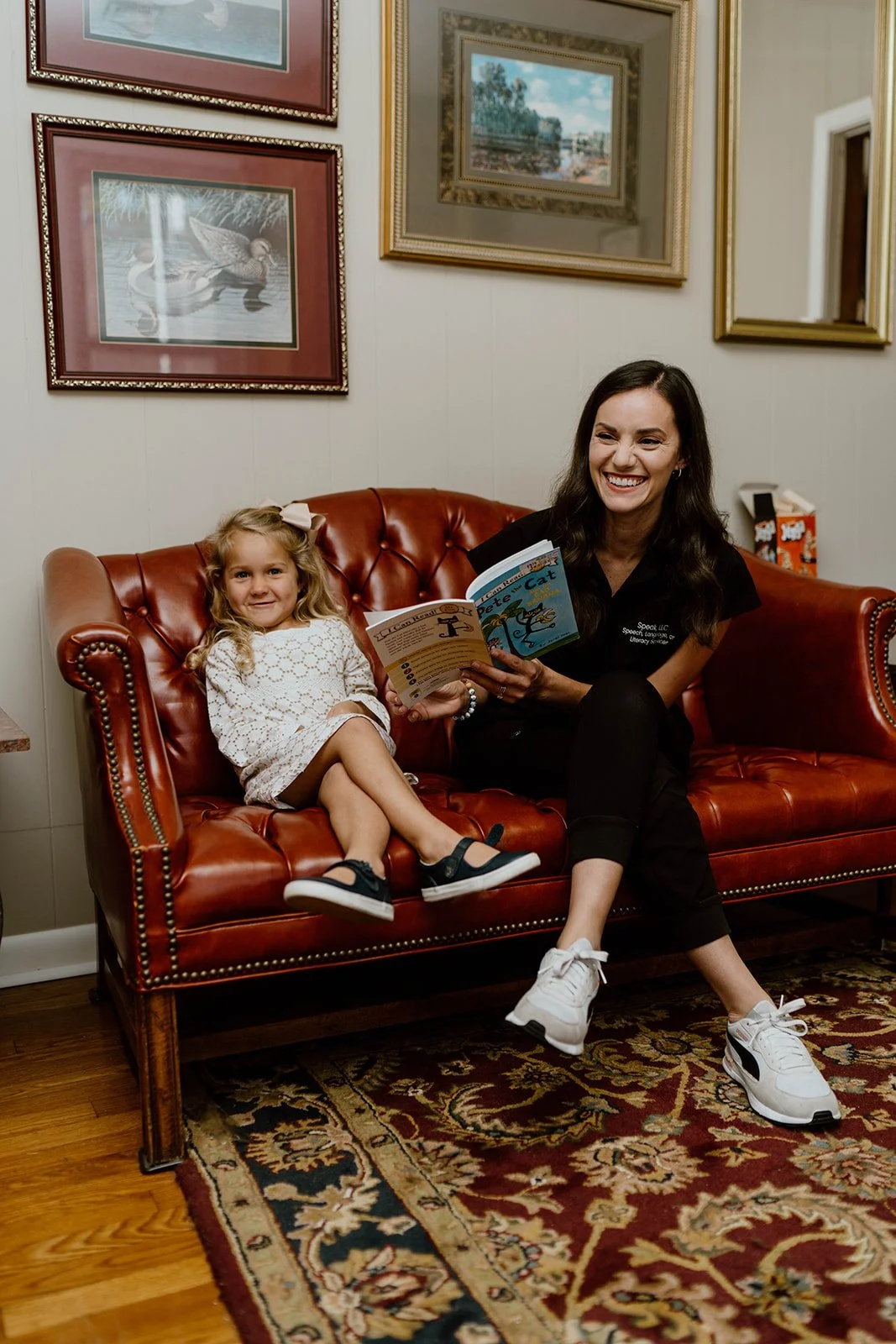A woman and a girl sitting on a red leather couch, reading a children's book titled 'Pete the Cat.' They are smiling, and the room has framed artwork on the walls and a patterned rug.