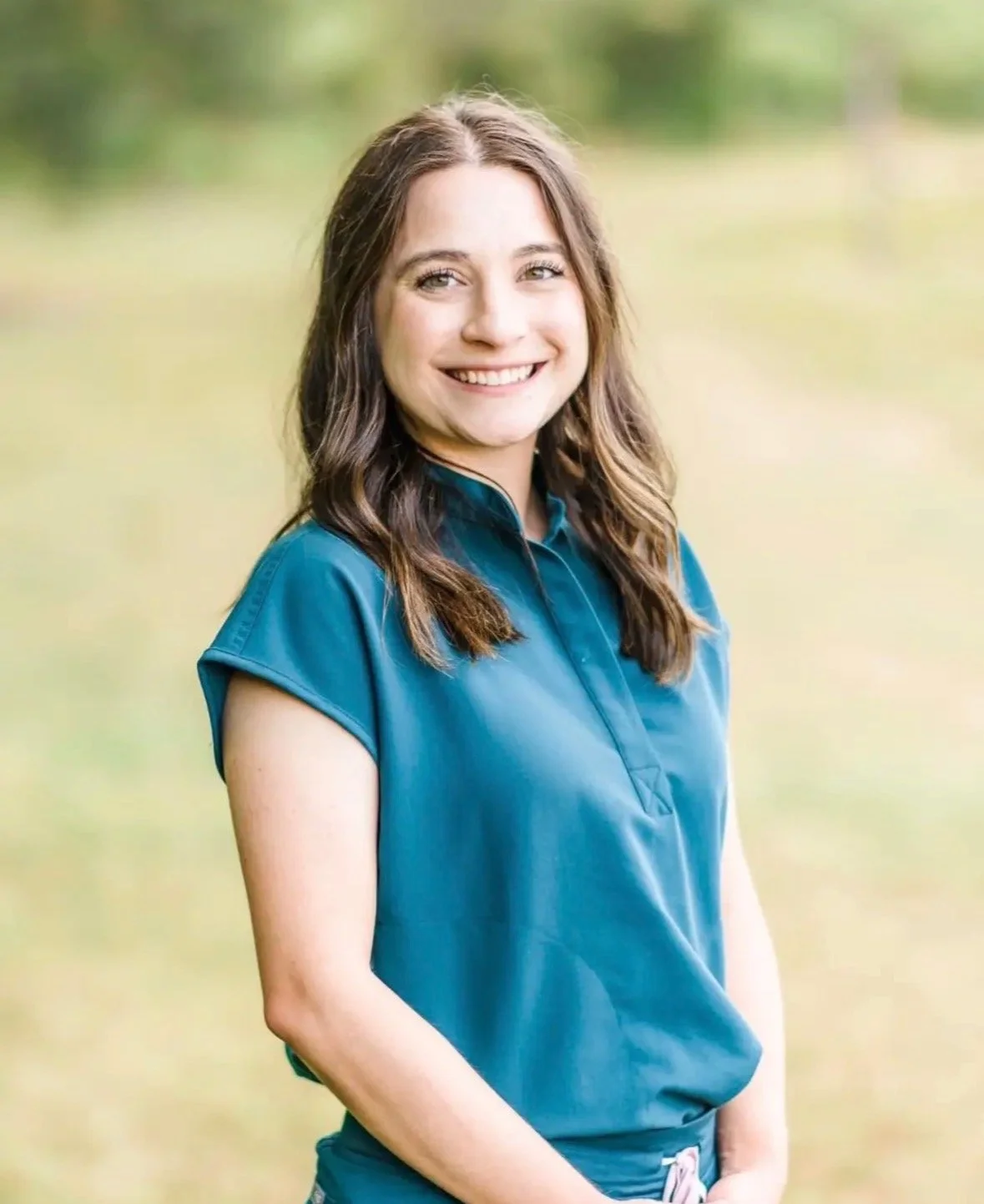 A young woman with long, wavy brown hair smiling outdoors, wearing a teal short-sleeved collared shirt, standing in a grassy area with blurred trees in the background.