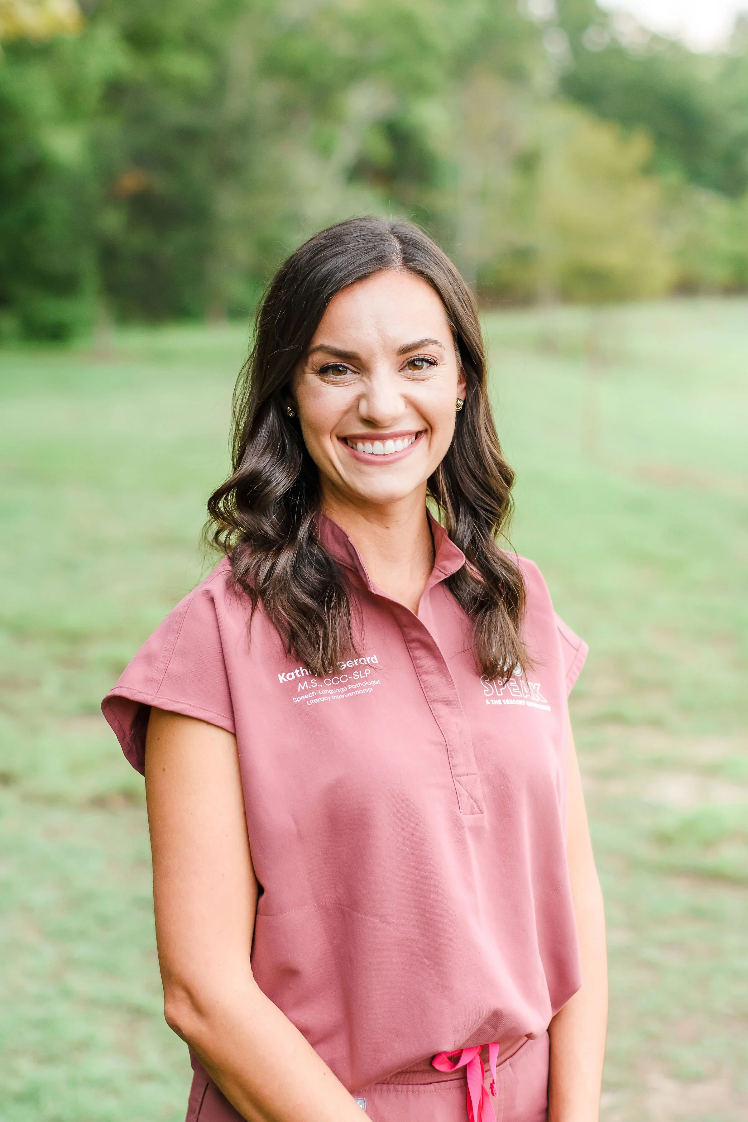 A smiling woman with long dark wavy hair, wearing a pink uniform shirt with embroidered text, standing outdoors in a blurred green park background.