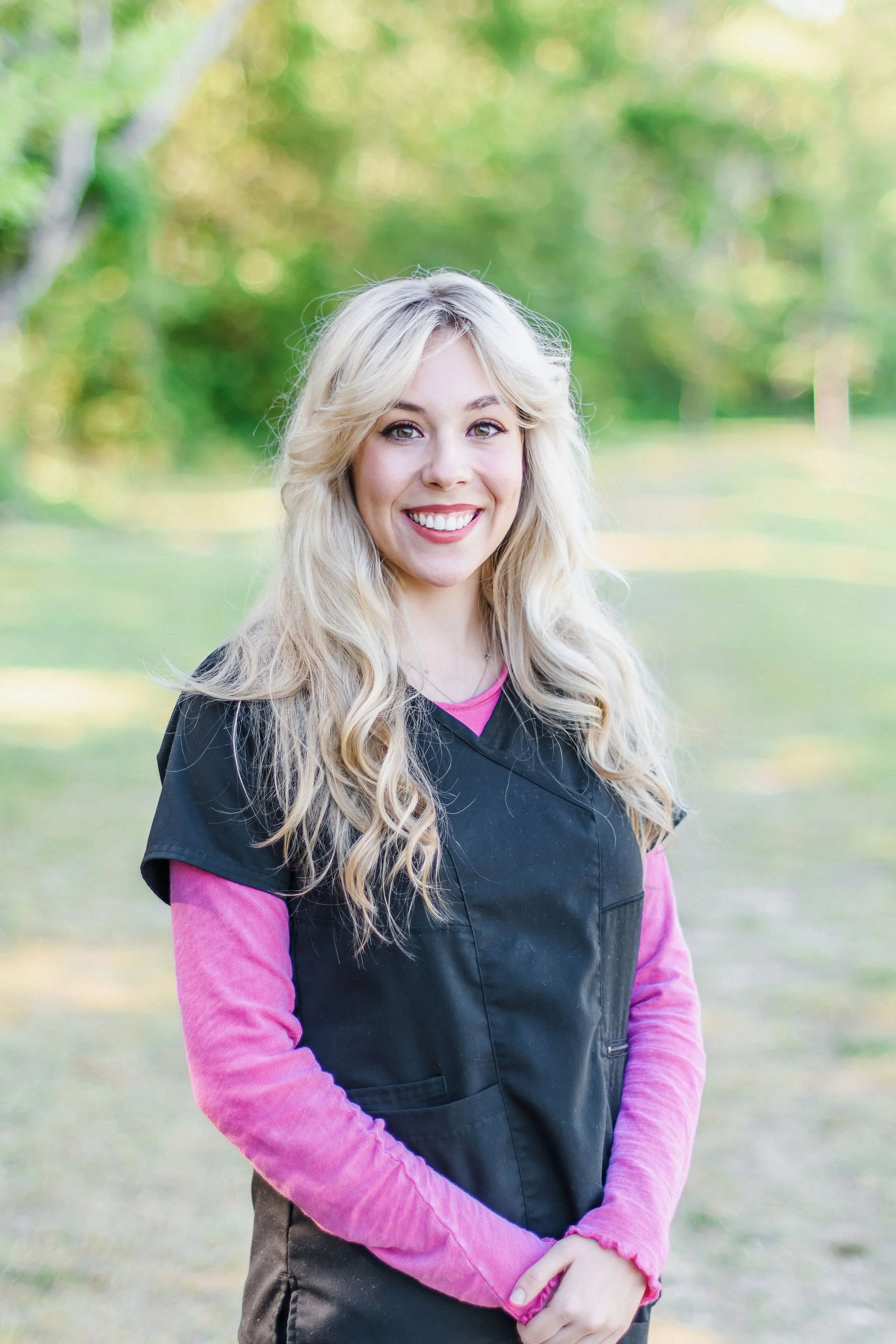 A young woman with long blonde hair and a bright smile, wearing a pink long-sleeve shirt under a black vest, standing outdoors in a park with green trees in the background.