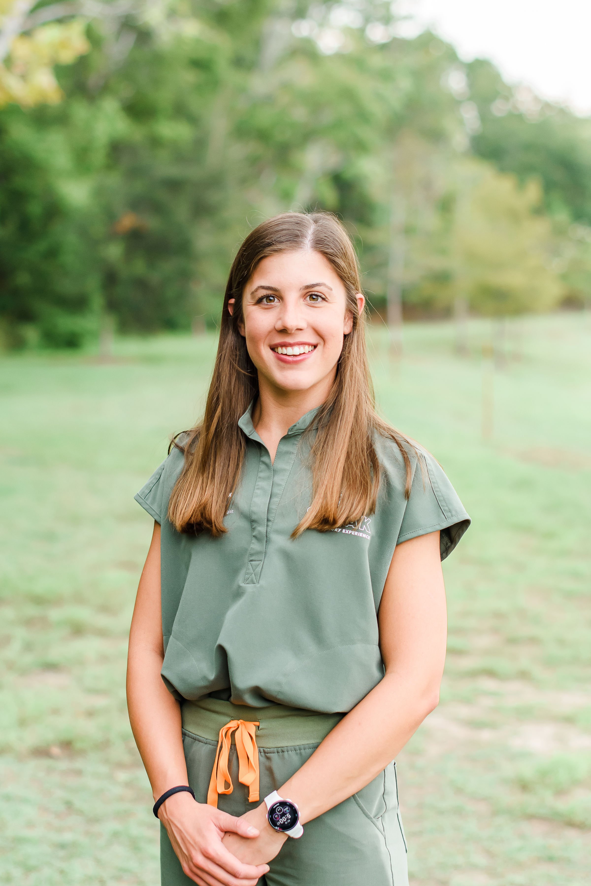 A young woman with long brown hair standing outdoors in a park or field with green trees and grass in the background. She is smiling and wearing a green short-sleeved shirt, a black wristband, and a smartwatch.