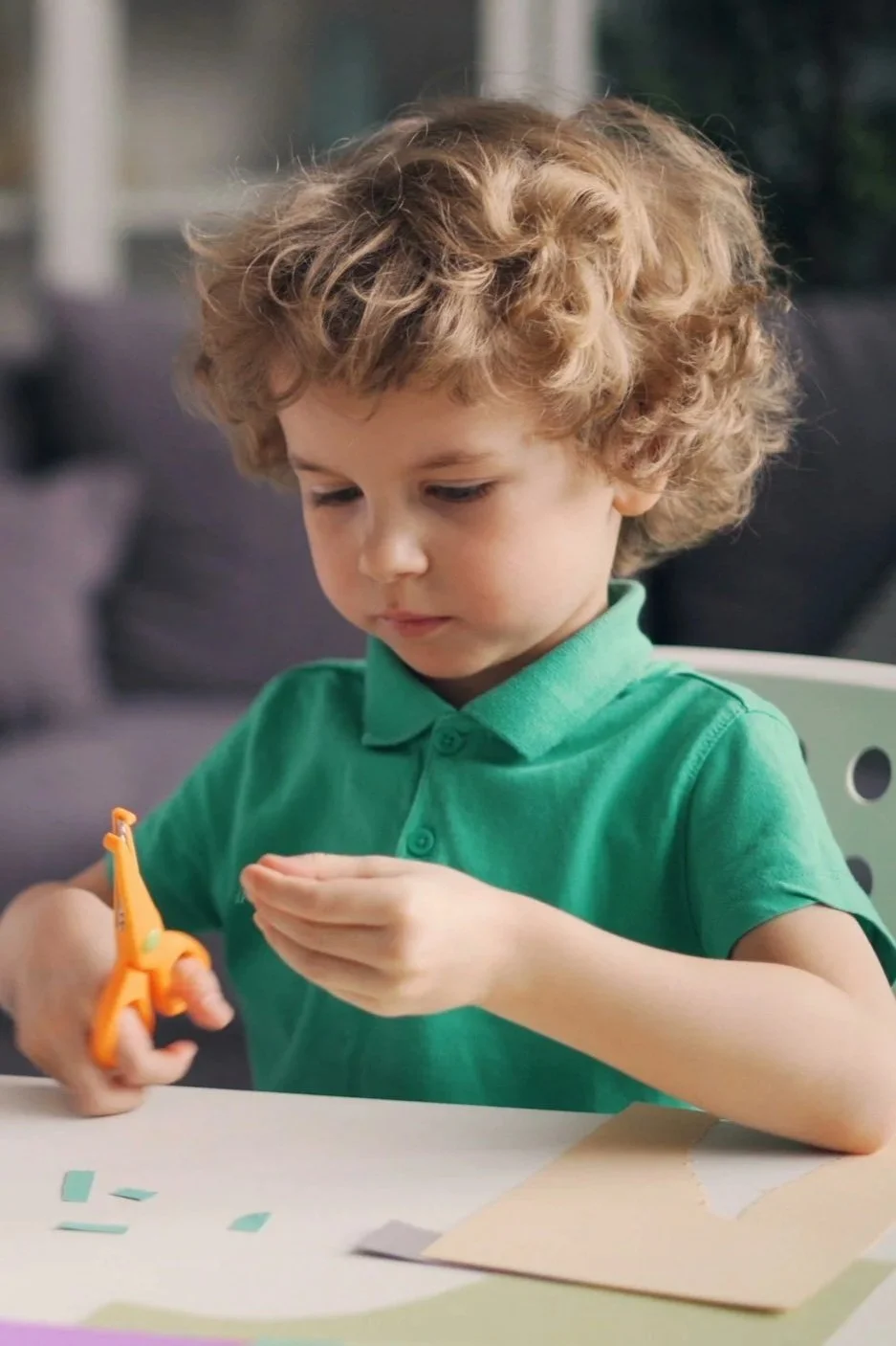 A young boy with curly blonde hair wearing a green polo shirt, using orange scissors to cut paper.