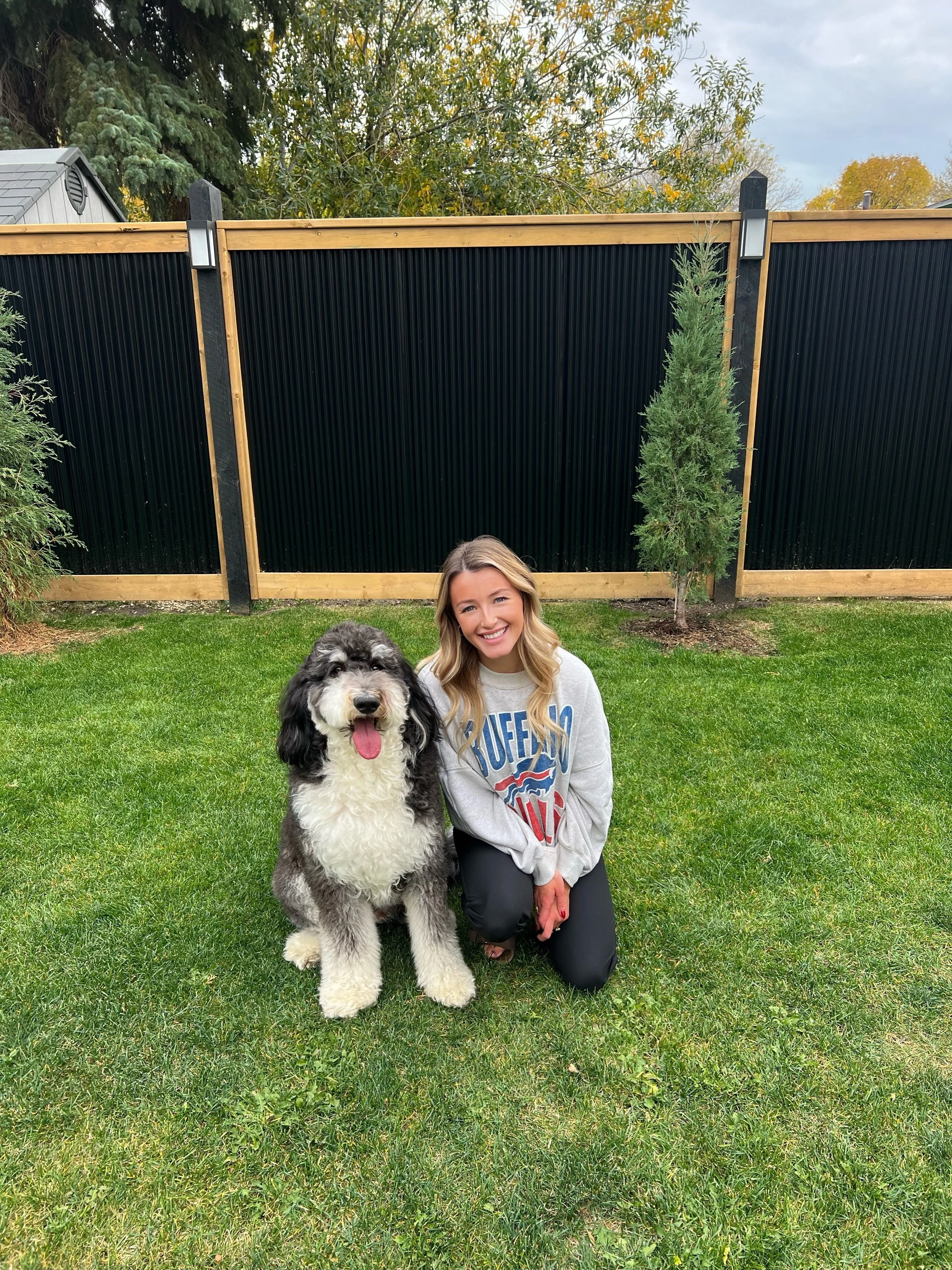 A woman with long blonde hair, smiling, kneeling on grass outdoors with a large fluffy black and white dog, possibly a Sheepadoodle, in front of a black fence surrounded by trees.