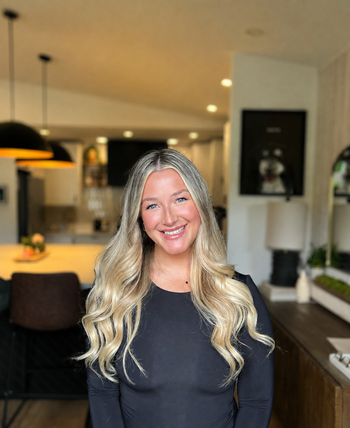 A woman with long blonde wavy hair smiling in a cozy modern home or office setting.