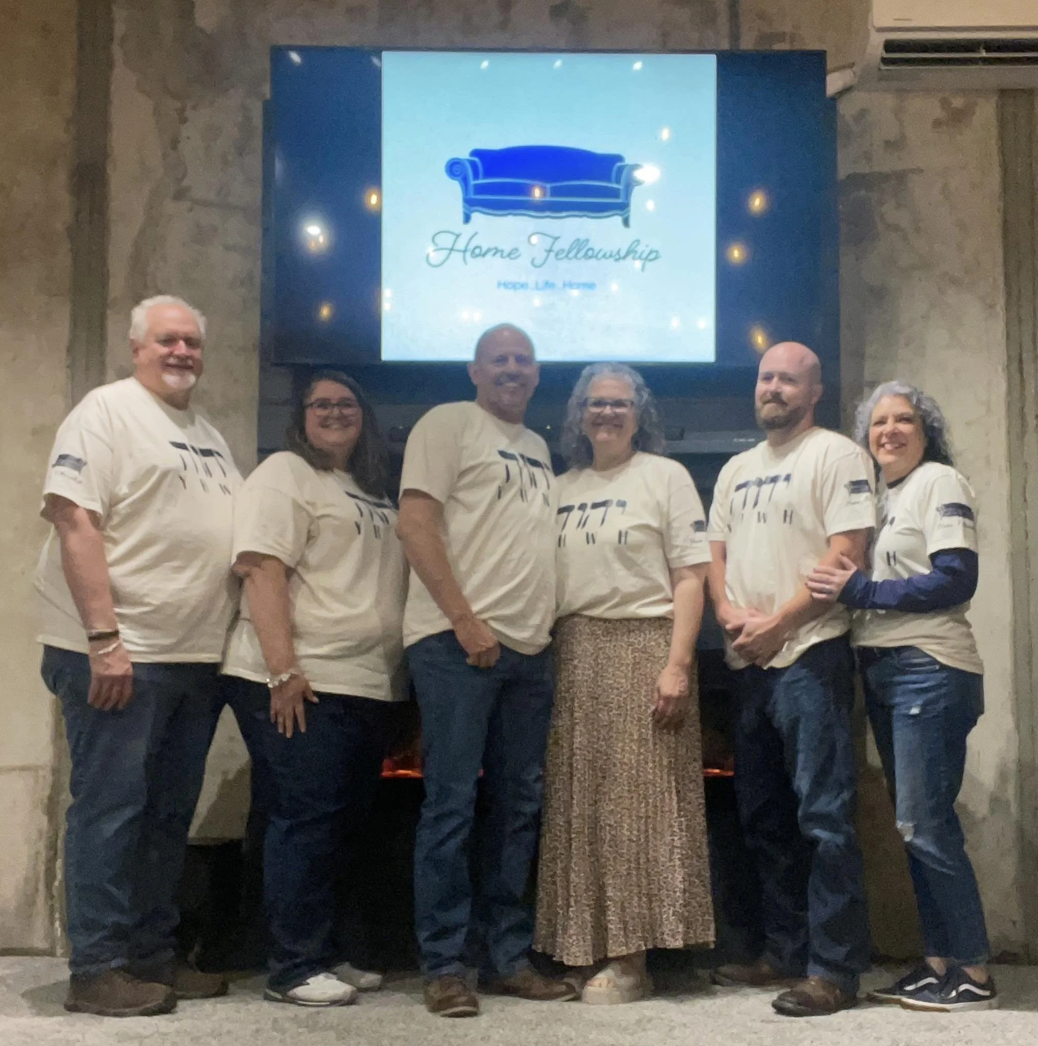Group of six diverse people standing together indoors, smiling, with a large screen behind them displaying a logo that features a blue couch and the words "Home Fellowship".