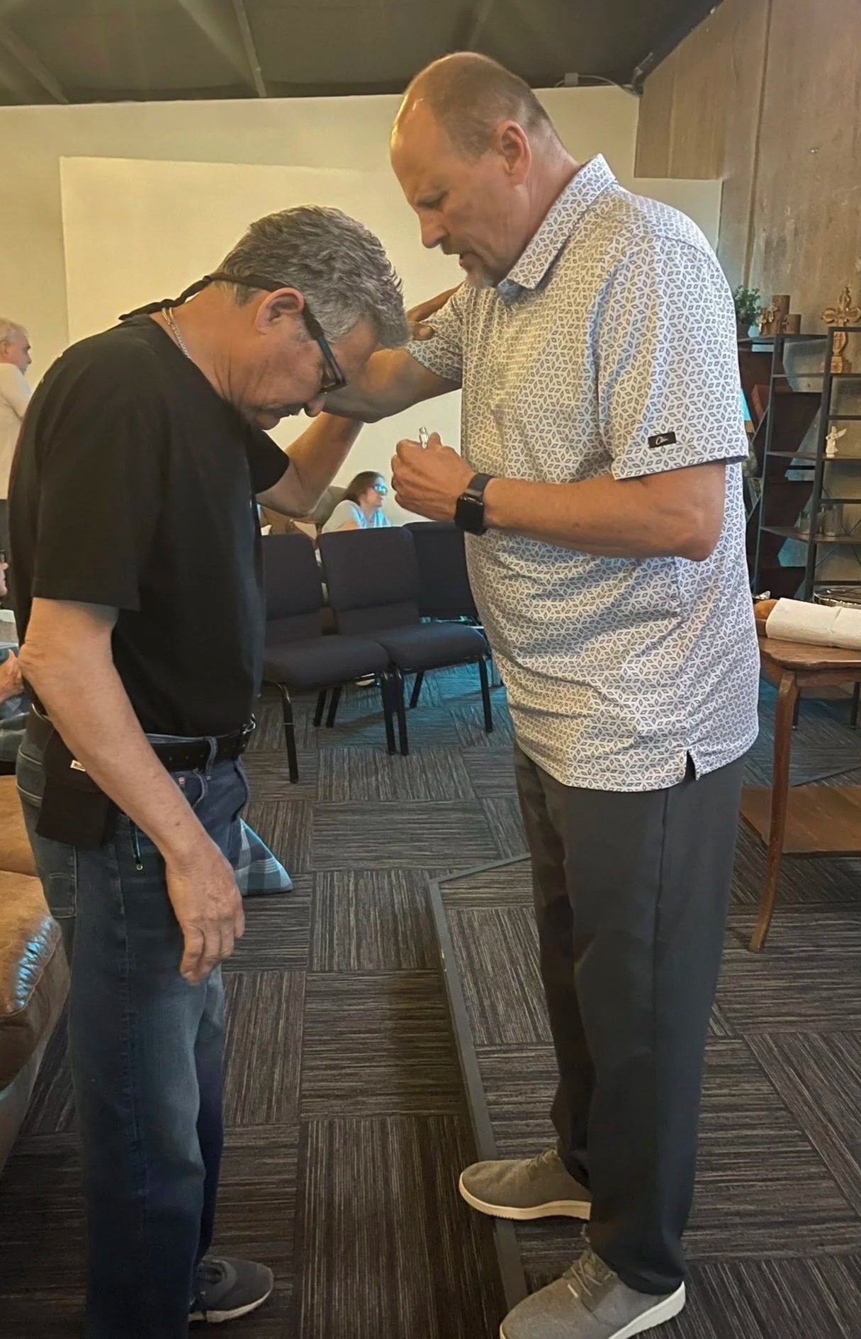 Two men standing indoors, bowing their heads and touching foreheads, with one man’s hand on the other's shoulder, in a moment of prayer or reflection.