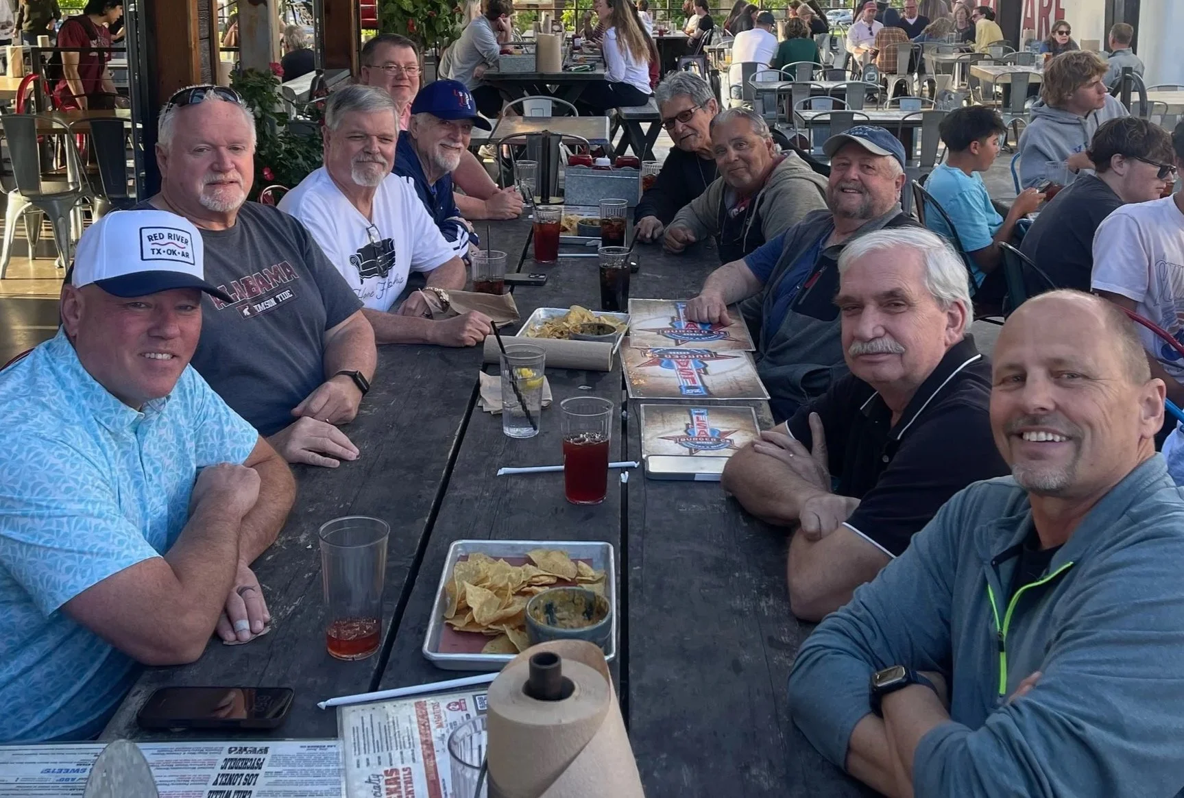 Group of men sitting at an outdoor restaurant table, enjoying food and drinks, with other diners visible in the background.