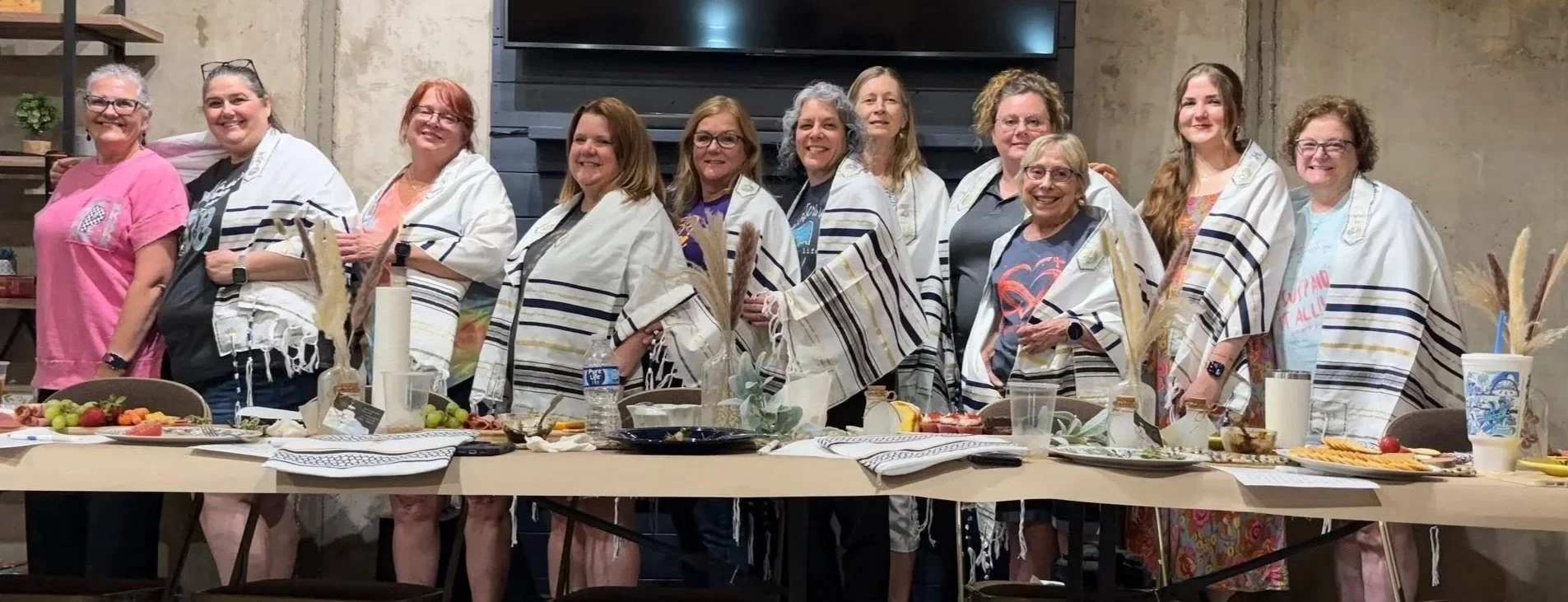 A group of women standing behind a table with various food items, all wearing white and black striped prayer shawls, in an indoor setting with a concrete wall and shelving in the background.