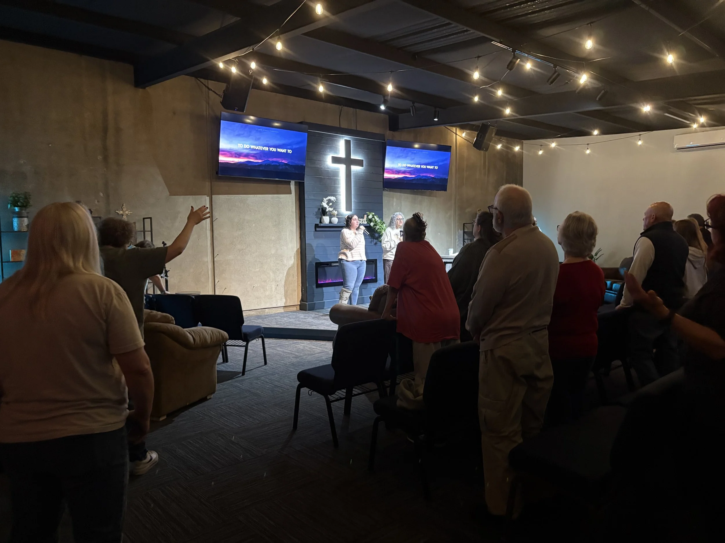 People attending a church service or gathering, some standing and some sitting, with a cross and two screens displaying music lyrics or messages in the background, inside a room with string lights on the ceiling.