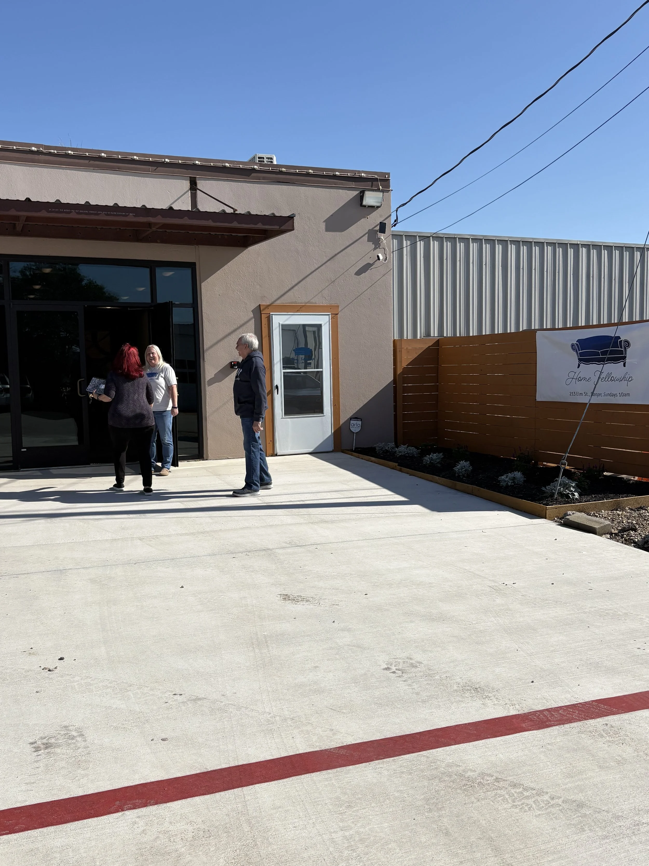 Three people standing outside a building, two women and one man, engaged in conversation. The building has a large glass door, a small window, and a sign reading 'Home Fellowship' on a wooden fence. The sky is clear and blue, with some electrical wires overhead.