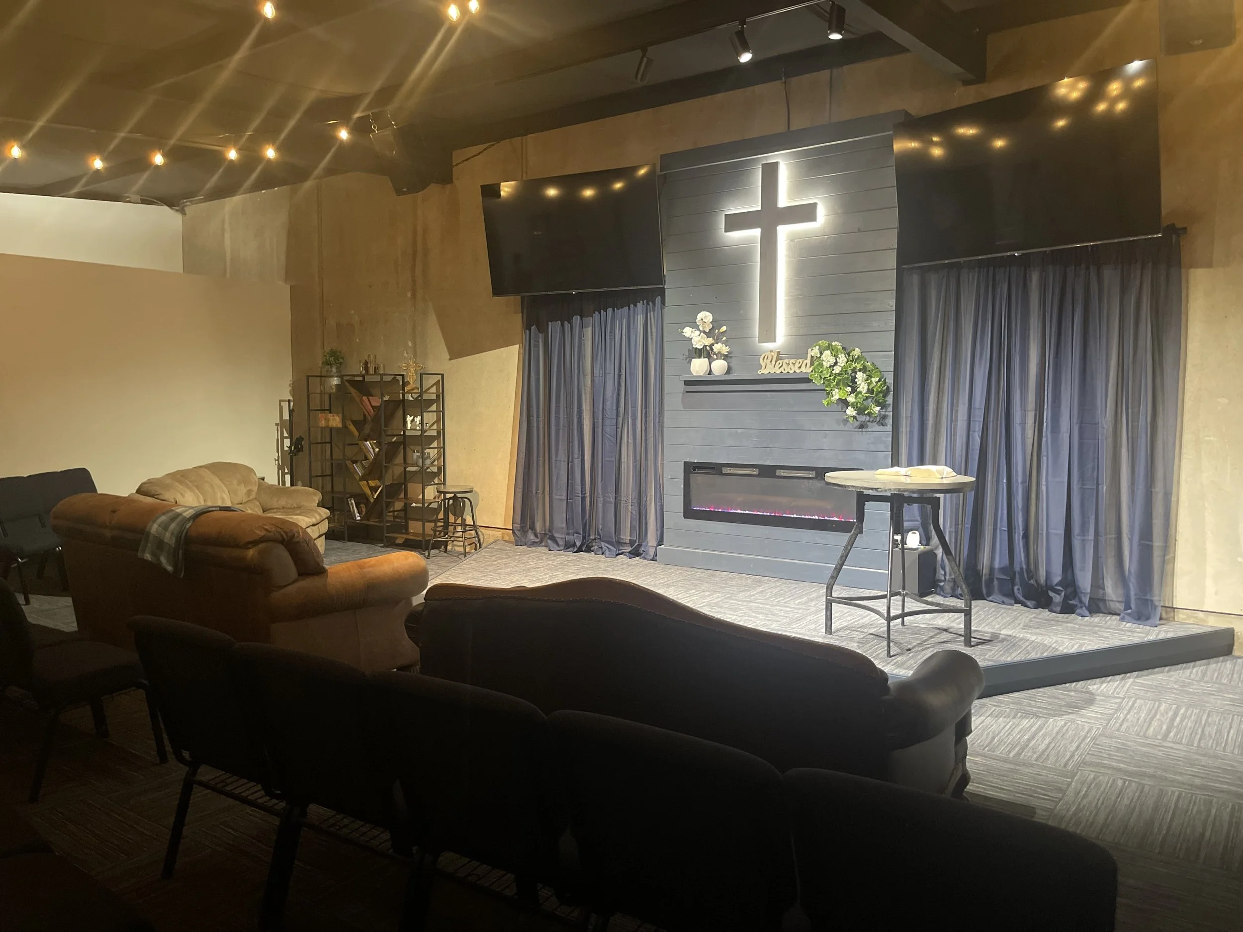 Interior view of a church or worship space with a prominent cross on a gray wooden wall, surrounded by blue curtains, flowers, and a sign that reads 'Blessed.' There are couches and chairs in the seating area, and string lights hanging from the ceiling.