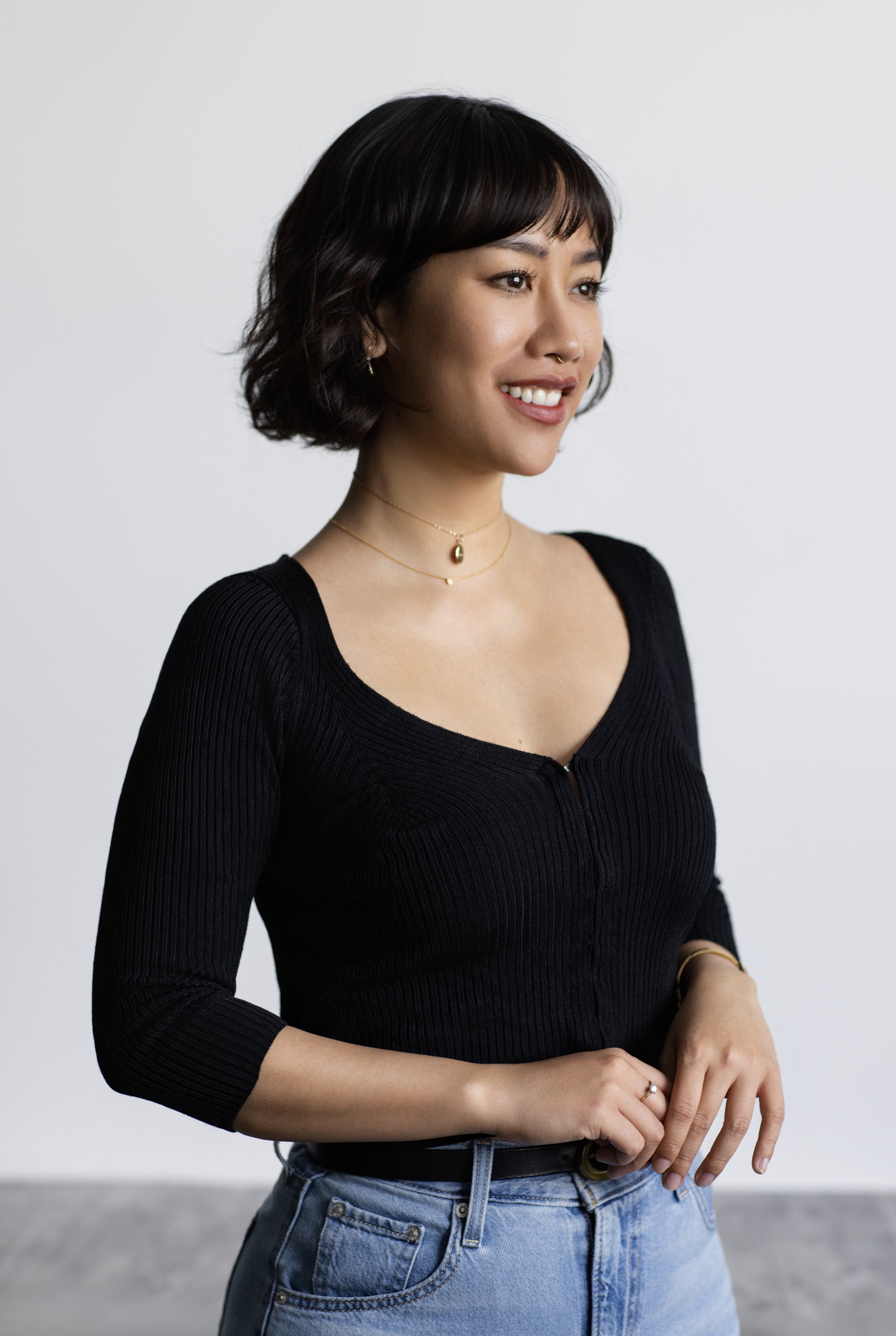 A woman with short dark hair, wearing a black ribbed long sleeve top and blue jeans, smiles slightly and faces to the right, standing against a plain white background.