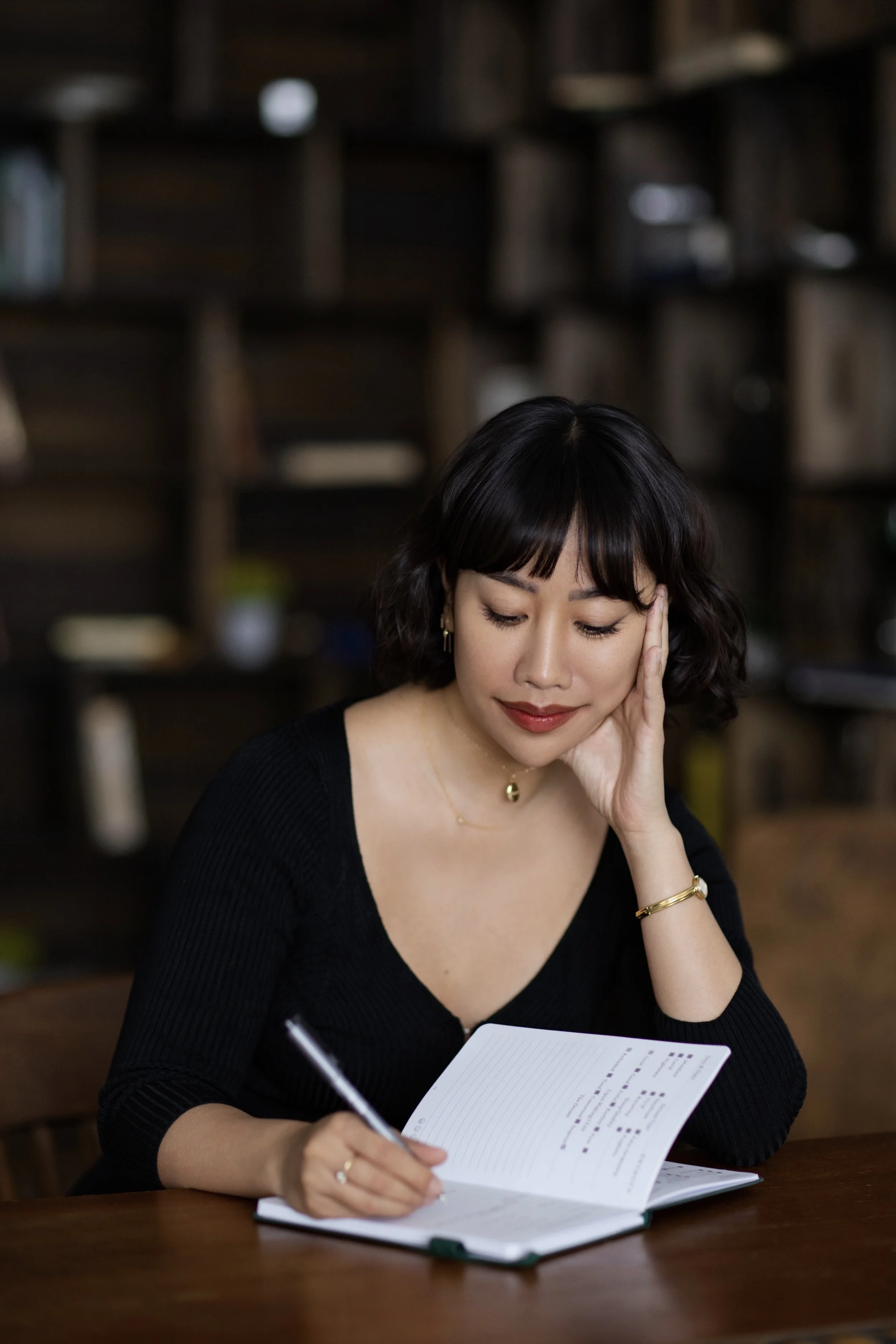 A woman with short dark hair and a black top sitting at a wooden table, holding a pen and writing in a notebook. She is resting her head on her left hand and appears to be focused or thoughtful. The background features a dark wooden shelf with various books and decorative items.