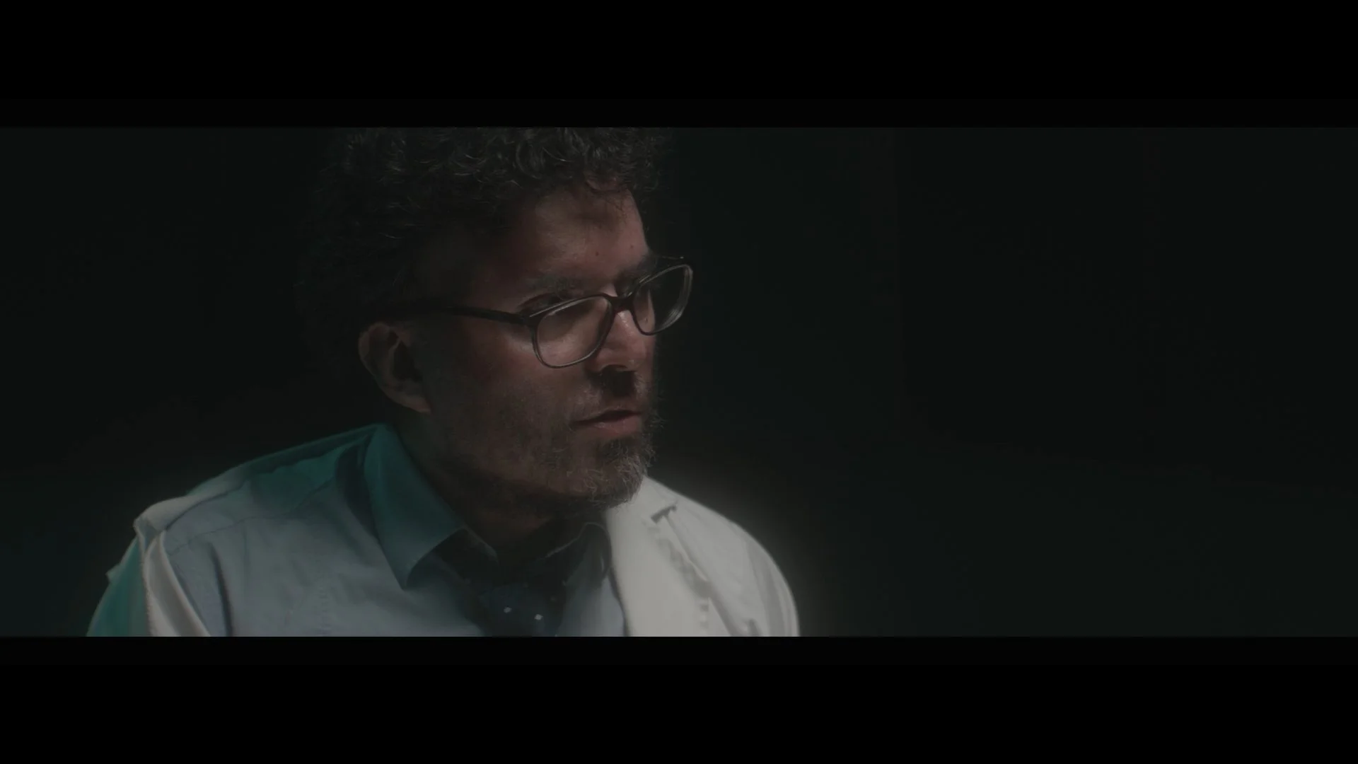 A man with curly hair and glasses, wearing a white shirt and tie, appears to be in a dark room.