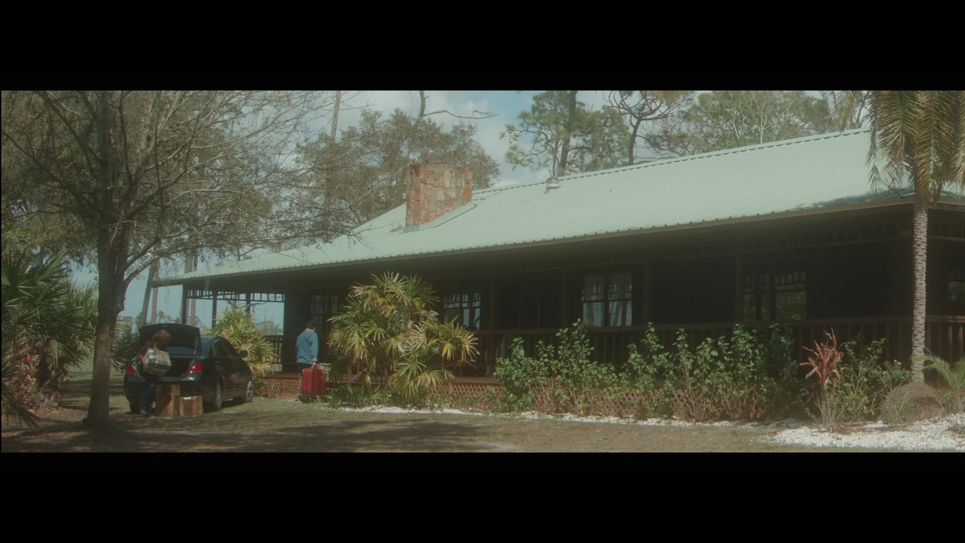 A house with a metal roof surrounded by trees and plants. Two people are seen near an SUV with its trunk open, and one is pulling a suitcase.