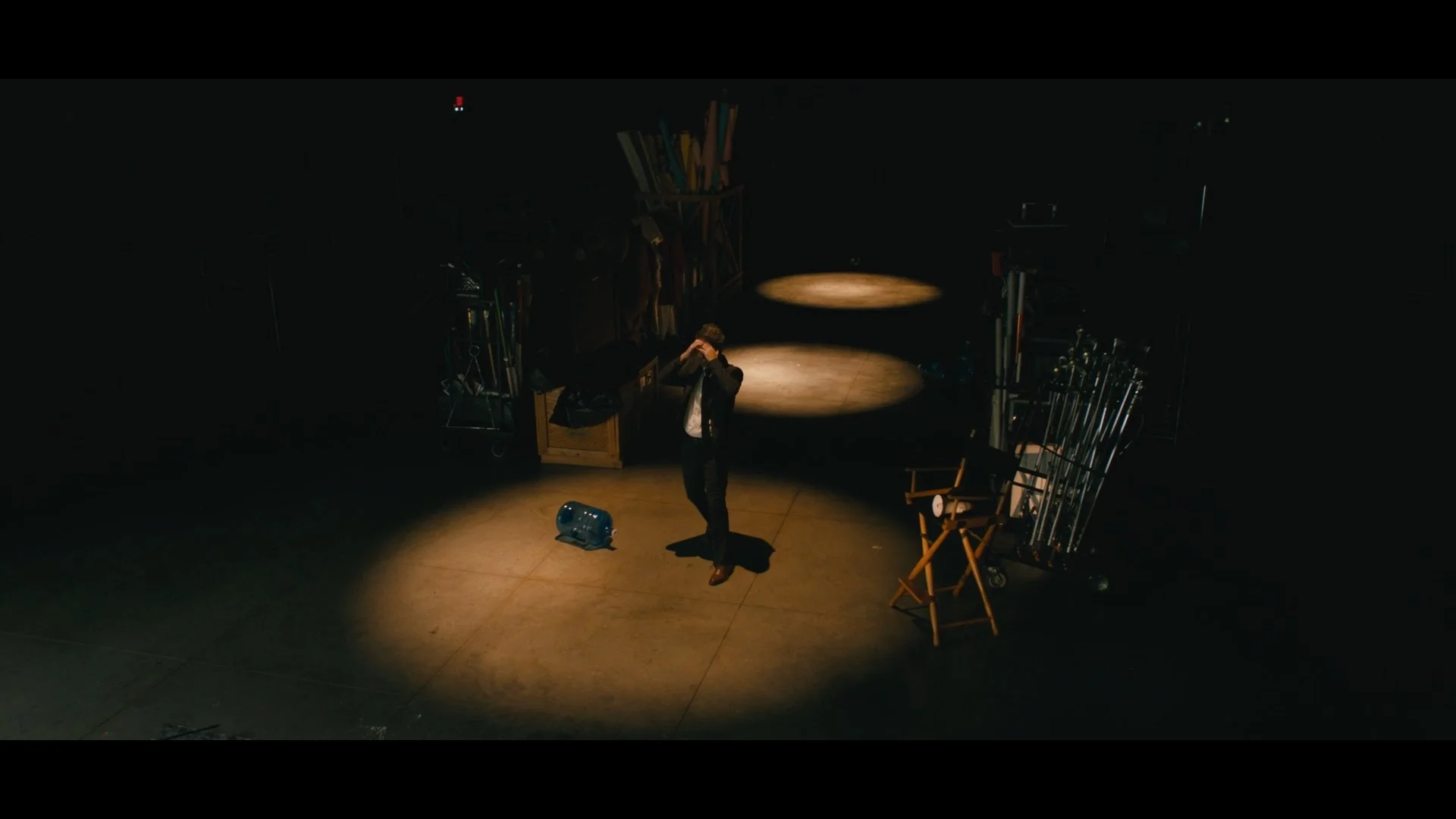 A person in a dark room standing under a spotlight, surrounded by various film equipment and tools, with their hands on their head.
