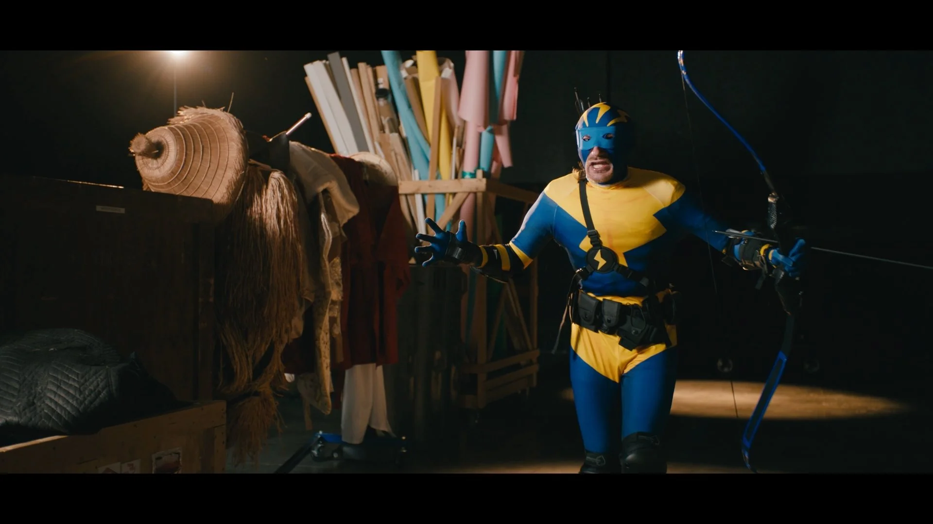A man dressed as a superhero in a colorful blue and yellow costume with a mask, standing in a room filled with various items like books, clothes, and a hay bale, holding a bow, and gesturing with his hand.