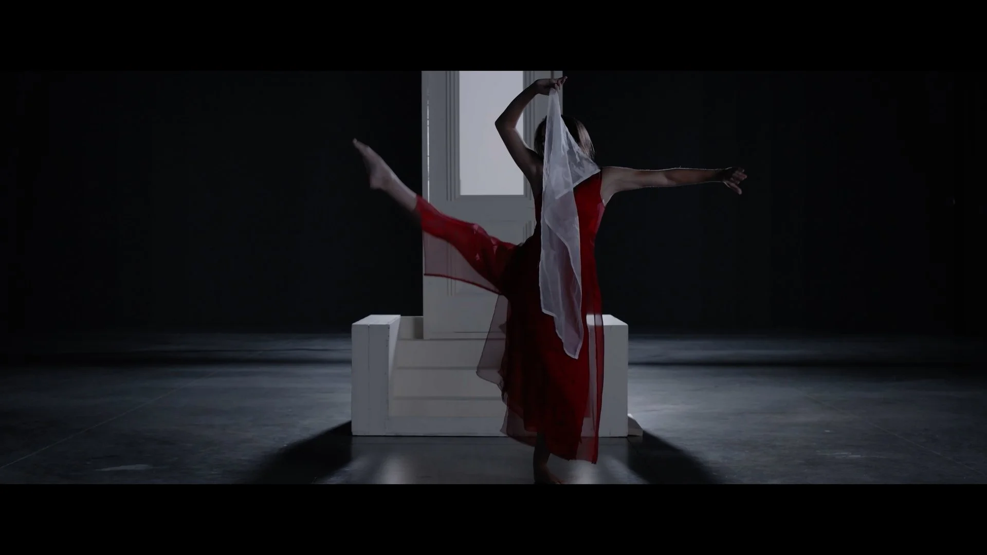 A dancer in a white and red dress performs a contemporary dance move in a dark, minimalistic room with a white rectangular platform and a white door in the background.