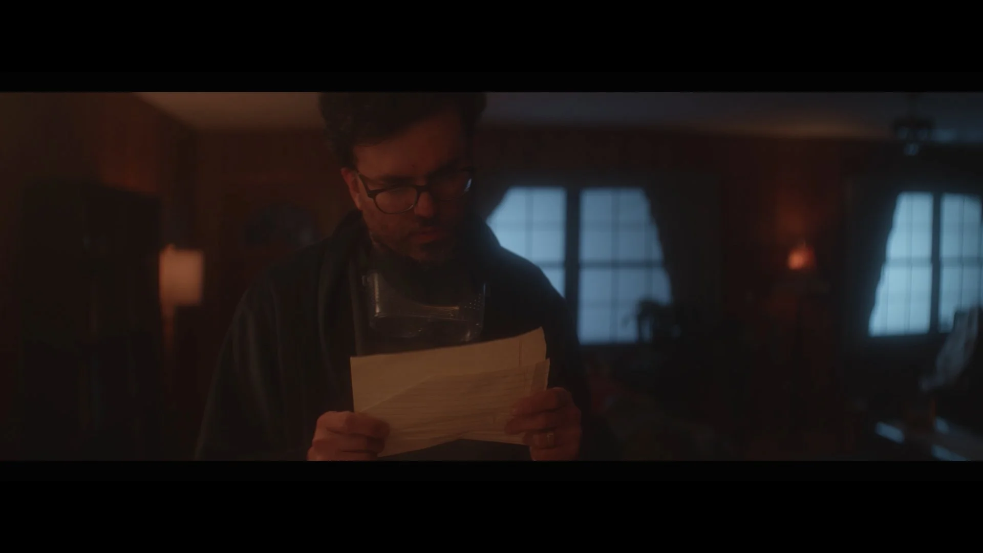 A man with glasses and a beard, wearing an apron and goggles around his neck, looks at a piece of paper in a dimly lit room with large windows and warm lighting.