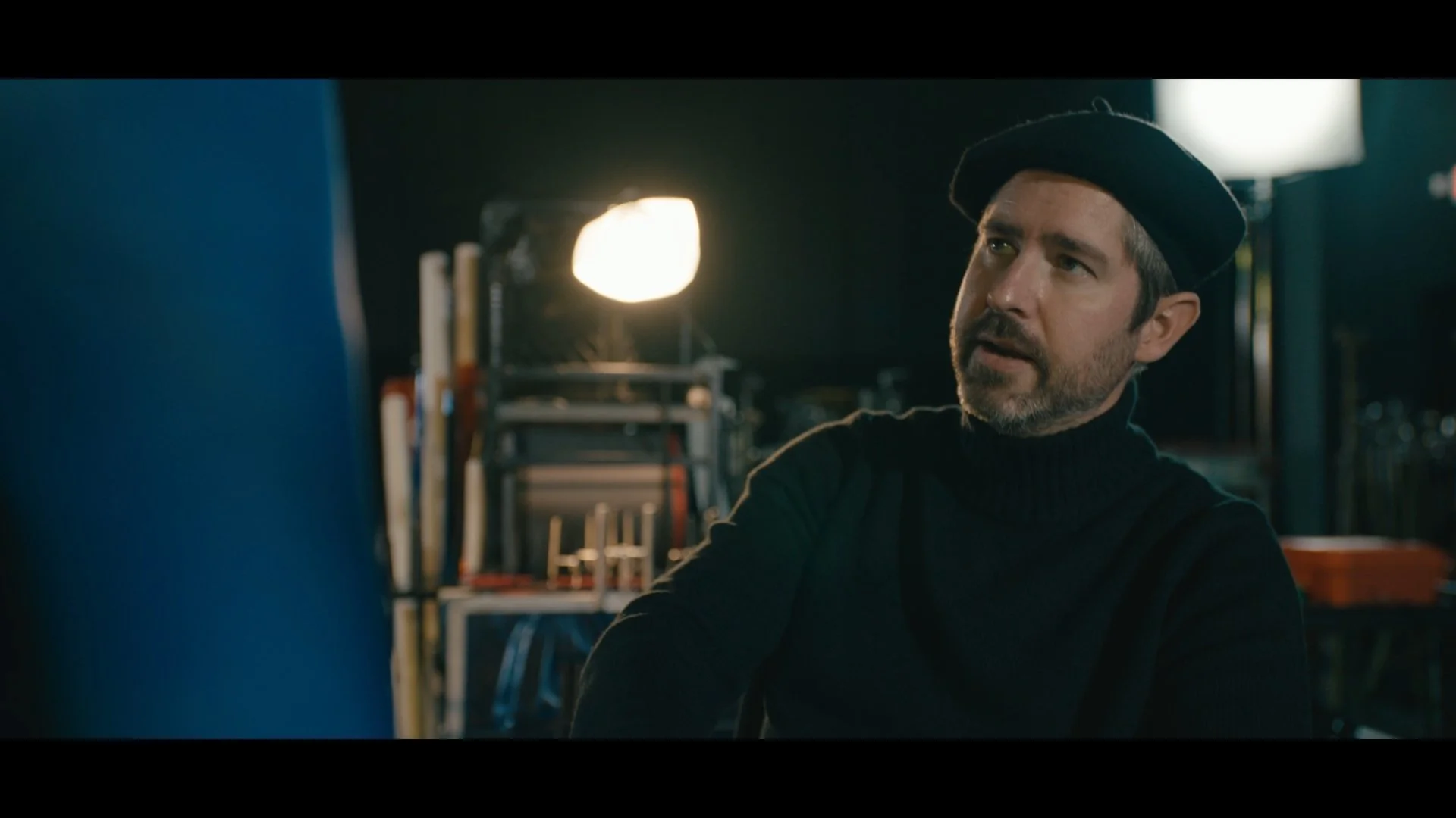 A man with a beard wearing a black beret and black turtleneck sitting in a dimly lit room with bookshelves and a bright lamp in the background.