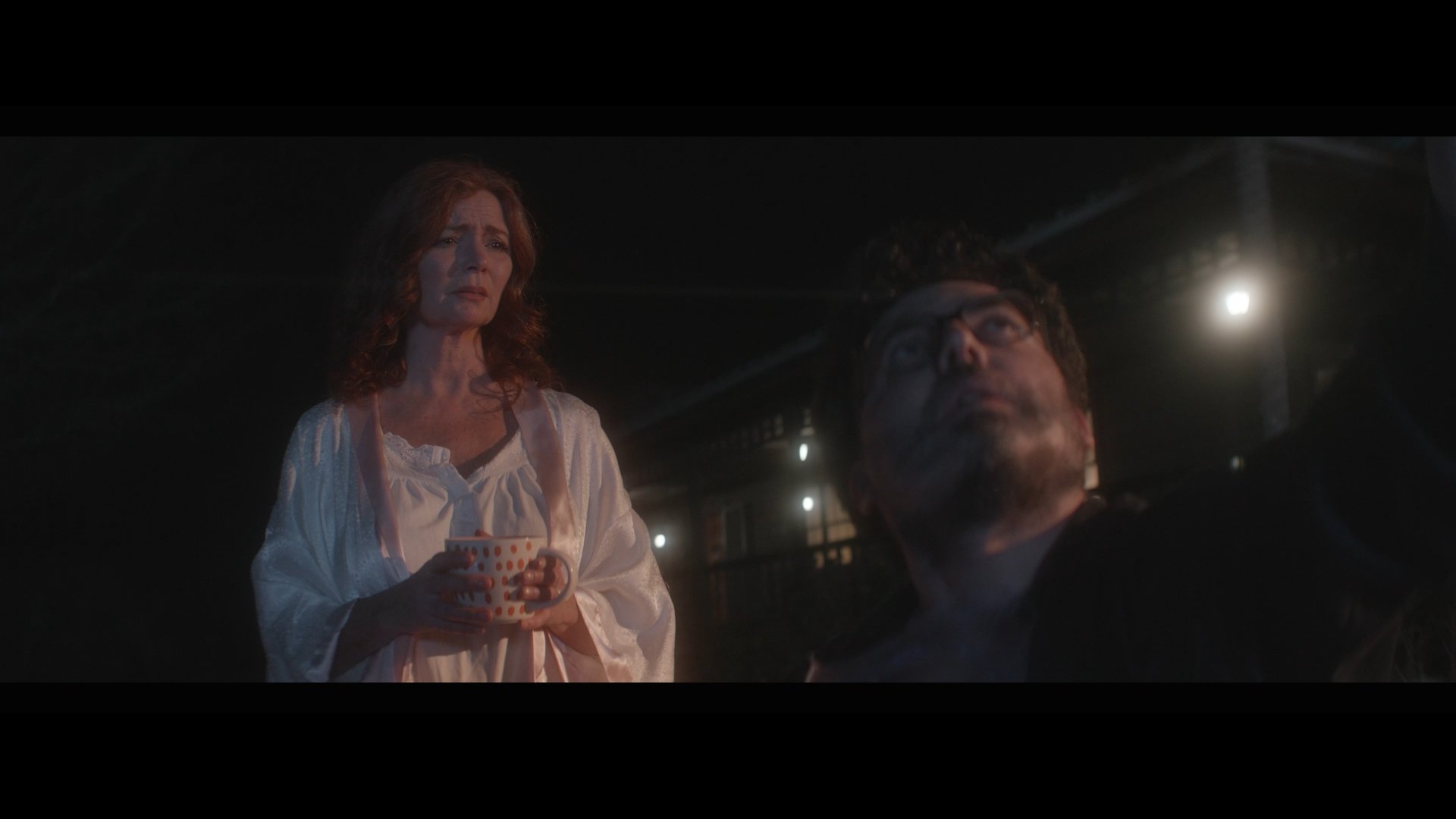 A woman holding a coffee mug stands behind a man sitting in a car at night, with the moon visible in the background.