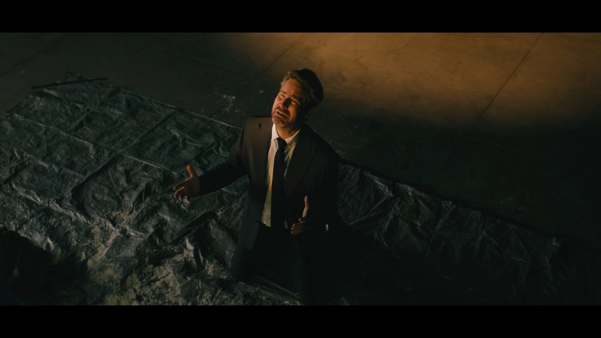 A man in a suit standing on a dark street at night, looking upward with a distressed expression, with a concrete wall and pavement visible.