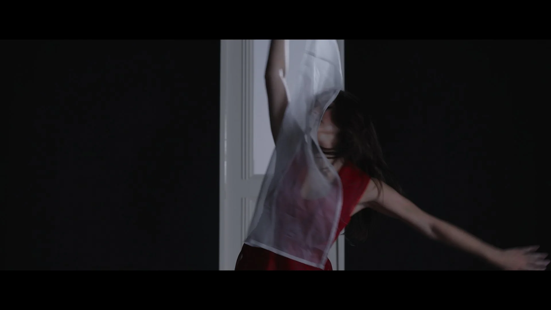 A woman with long dark hair in a red dress moves through a dark room, partially visible behind a door frame, with her arms extended.