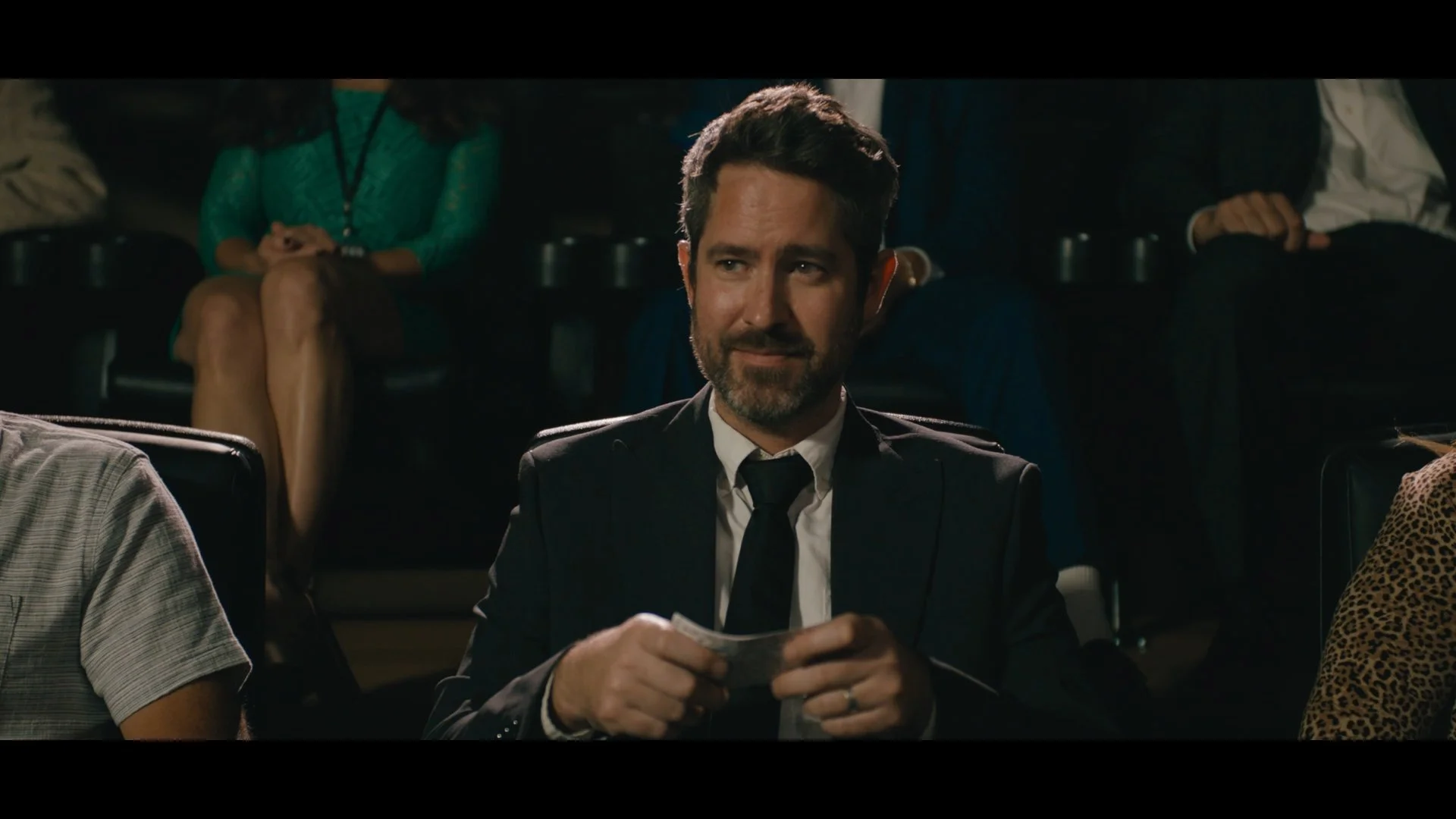 A man in a suit and tie is sitting in a theater, holding a ticket and smiling. Behind him, a woman in a green dress is sitting with her legs crossed, and other people are also seated in the background.